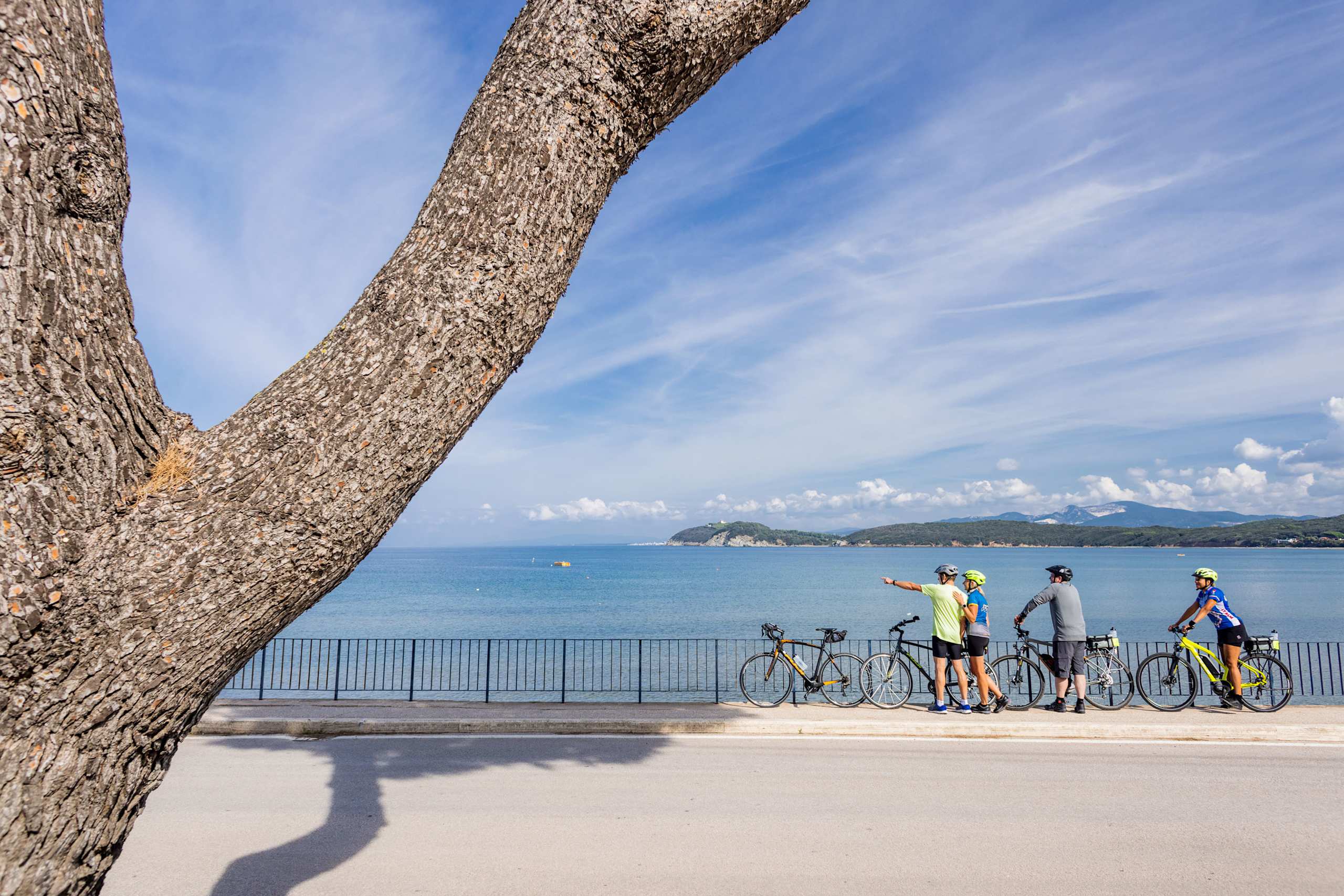 A group of cyclists stand on a beachfront promenade, with a scenic coastal landscape and blue sky visible in the background.