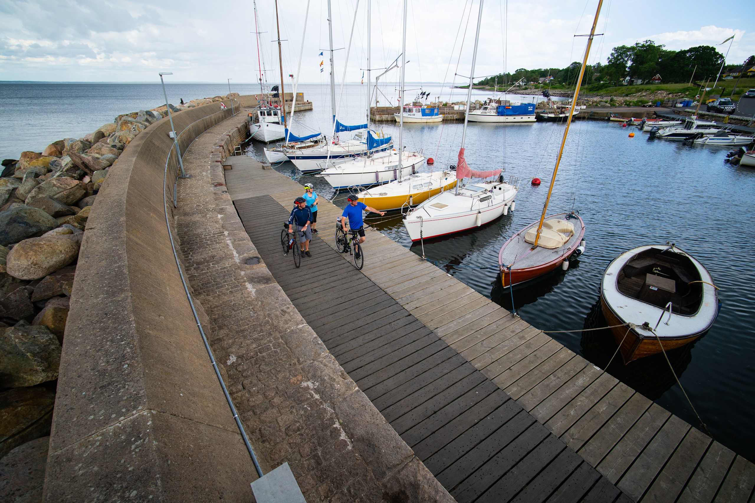 A wooden dock extends along the shoreline, with various sailboats and yachts moored in the calm waters of the harbor. In the background, a scenic landscape with trees and buildings can be seen.