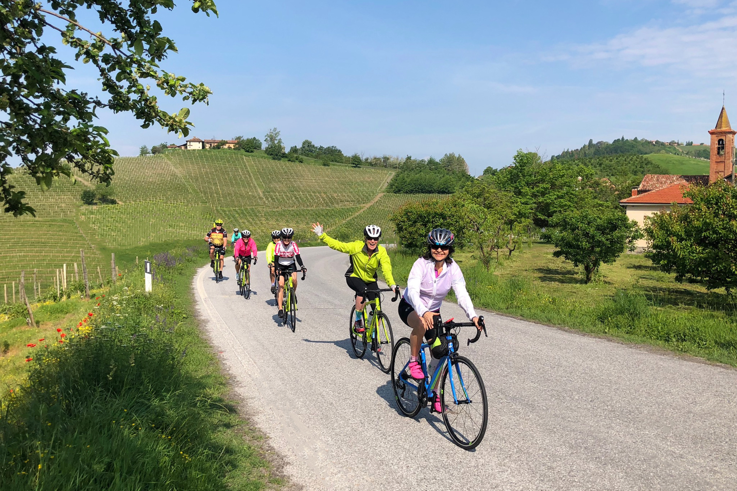 A group of cyclists riding on a gravel path surrounded by lush green hills, vineyards, and a small village in the distance.