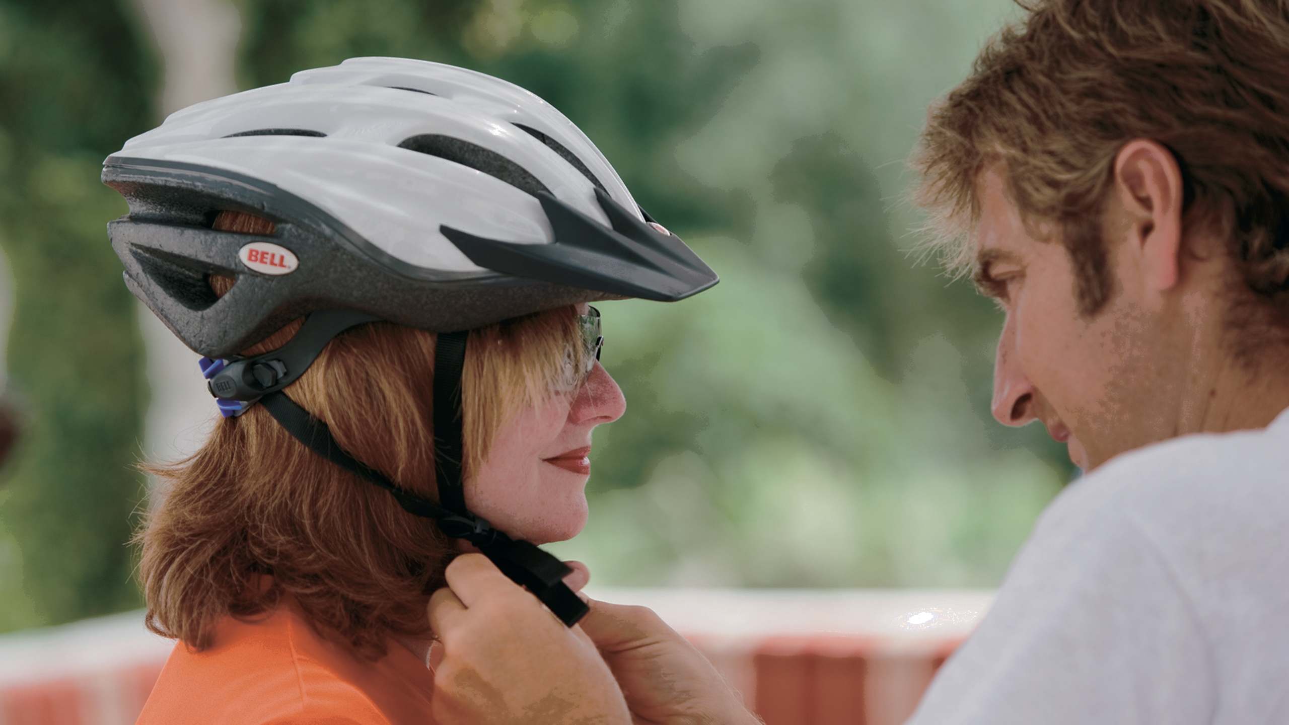 A young woman wearing a bicycle helmet is interacting with a man in an outdoor setting with greenery in the background.