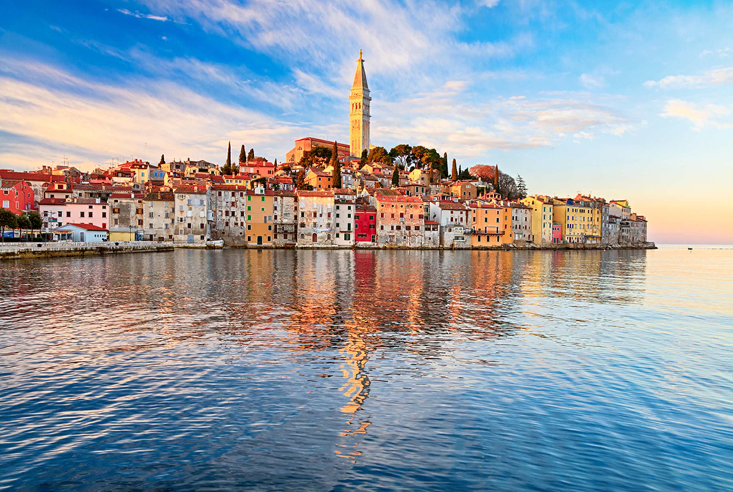 A picturesque coastal town with colorful buildings, a tall church spire, and a serene body of water reflecting the vibrant scene in the foreground.