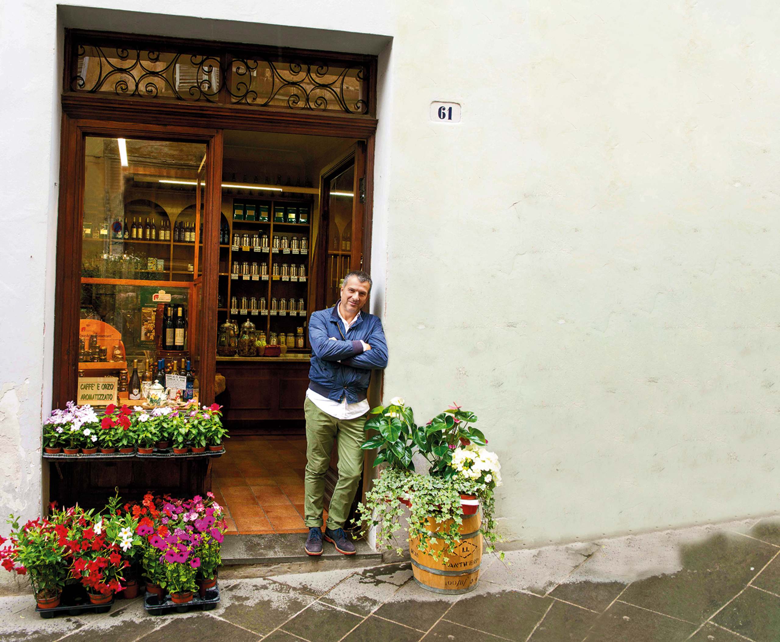 A person stands in the doorway of a quaint shop, surrounded by colorful flowers and a display of wine bottles and other goods inside the shop.