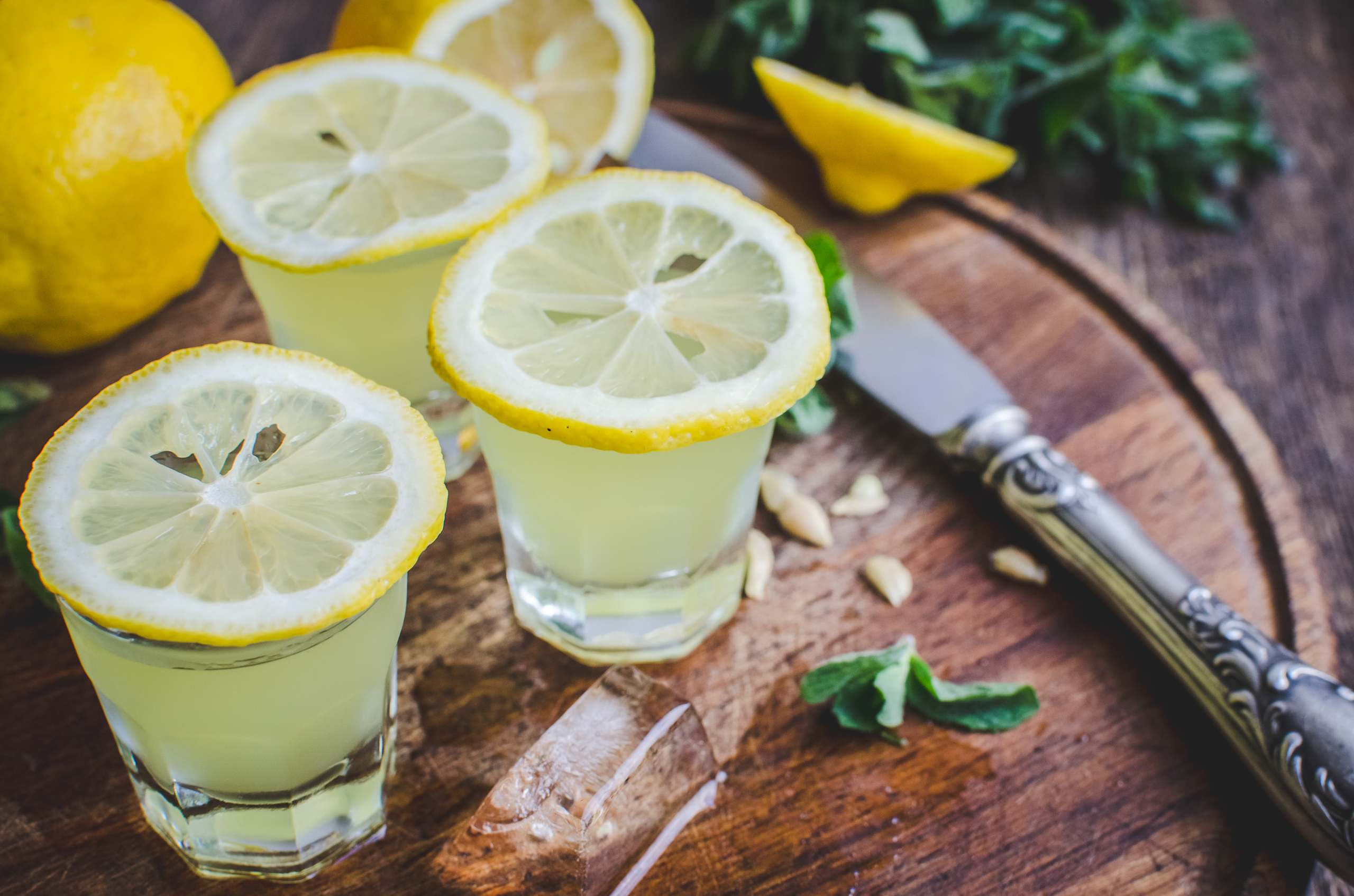 Two glasses filled with a bright yellow liquid, likely lemonade, sit on a wooden surface alongside fresh lemon slices and a pair of kitchen shears.
