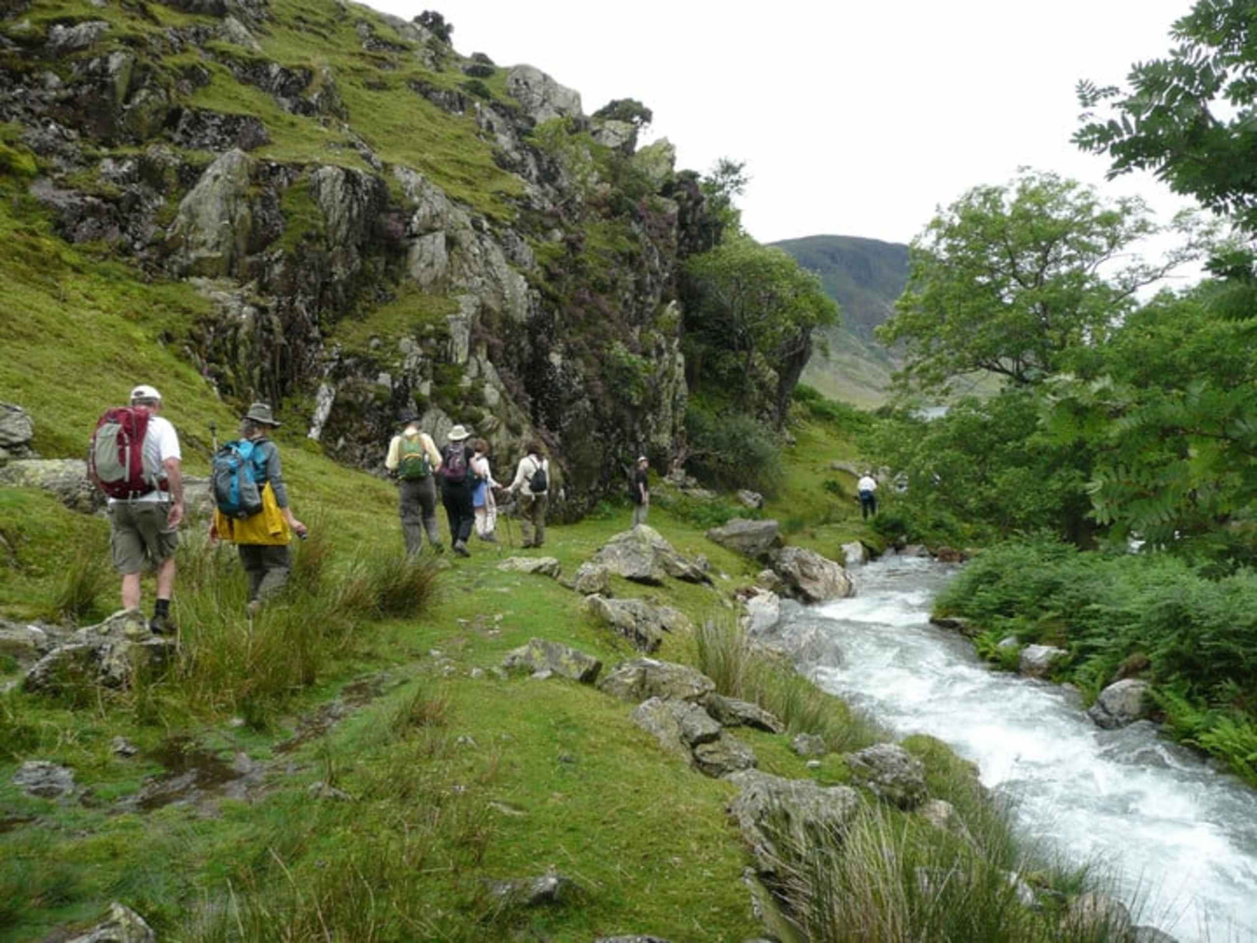 A group of hikers traversing a rocky, moss-covered trail alongside a rushing mountain stream, surrounded by lush, verdant vegetation and towering cliffs in the background.