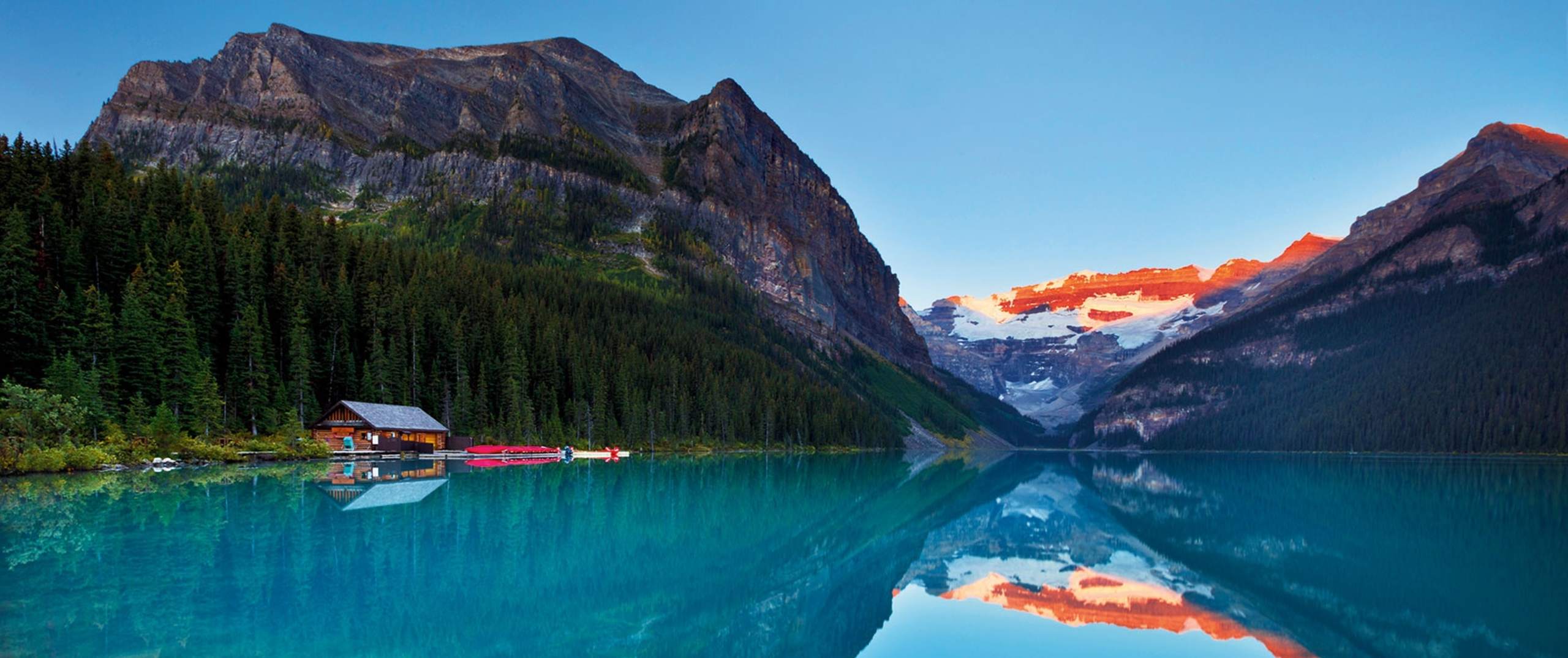 A serene alpine lake surrounded by majestic mountains, with a small wooden cabin reflected in the calm, turquoise waters, and the sky ablaze with vibrant colors at sunset.