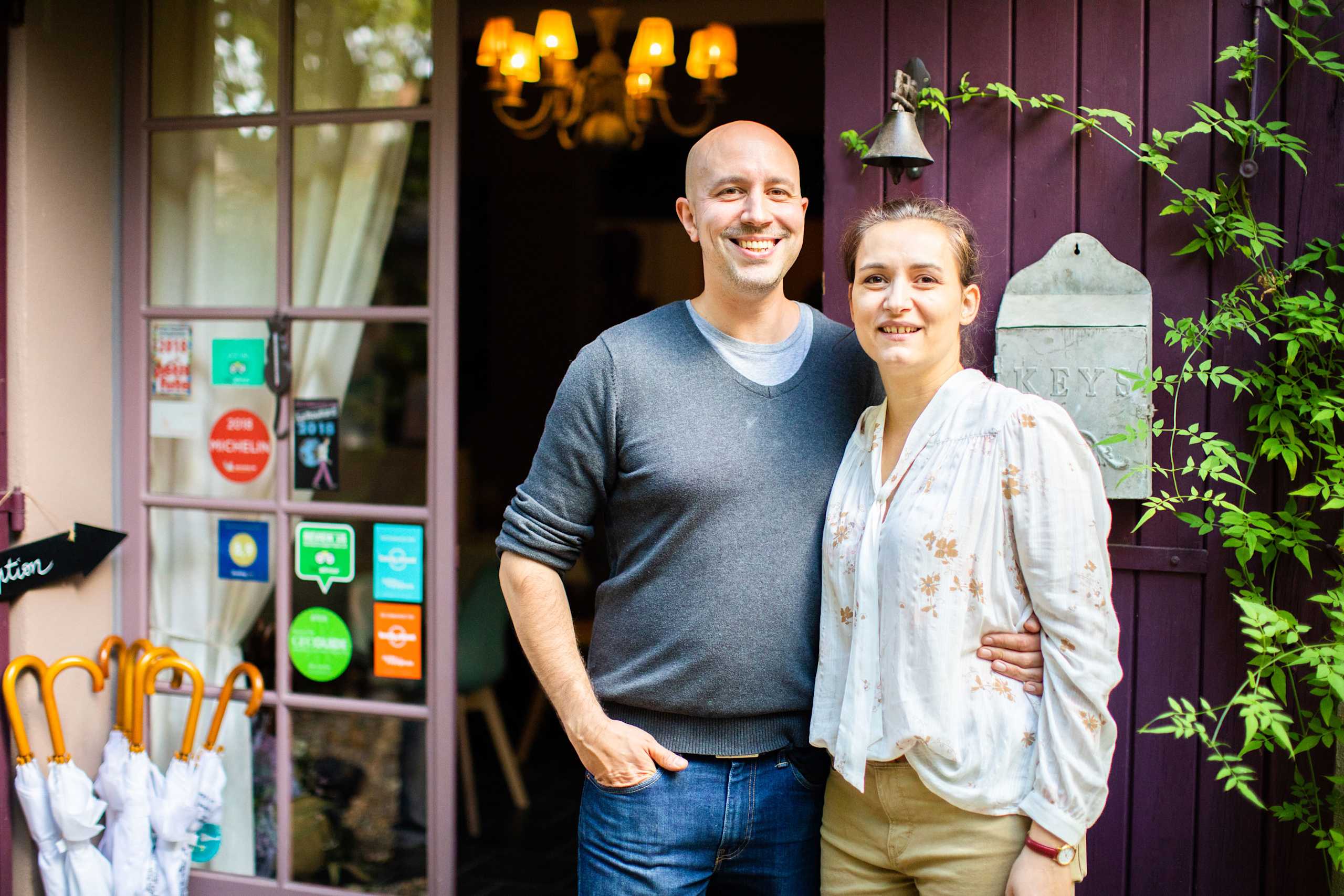 A smiling couple stands in front of a colorful, rustic wooden building, surrounded by lush greenery and a cozy, inviting atmosphere.