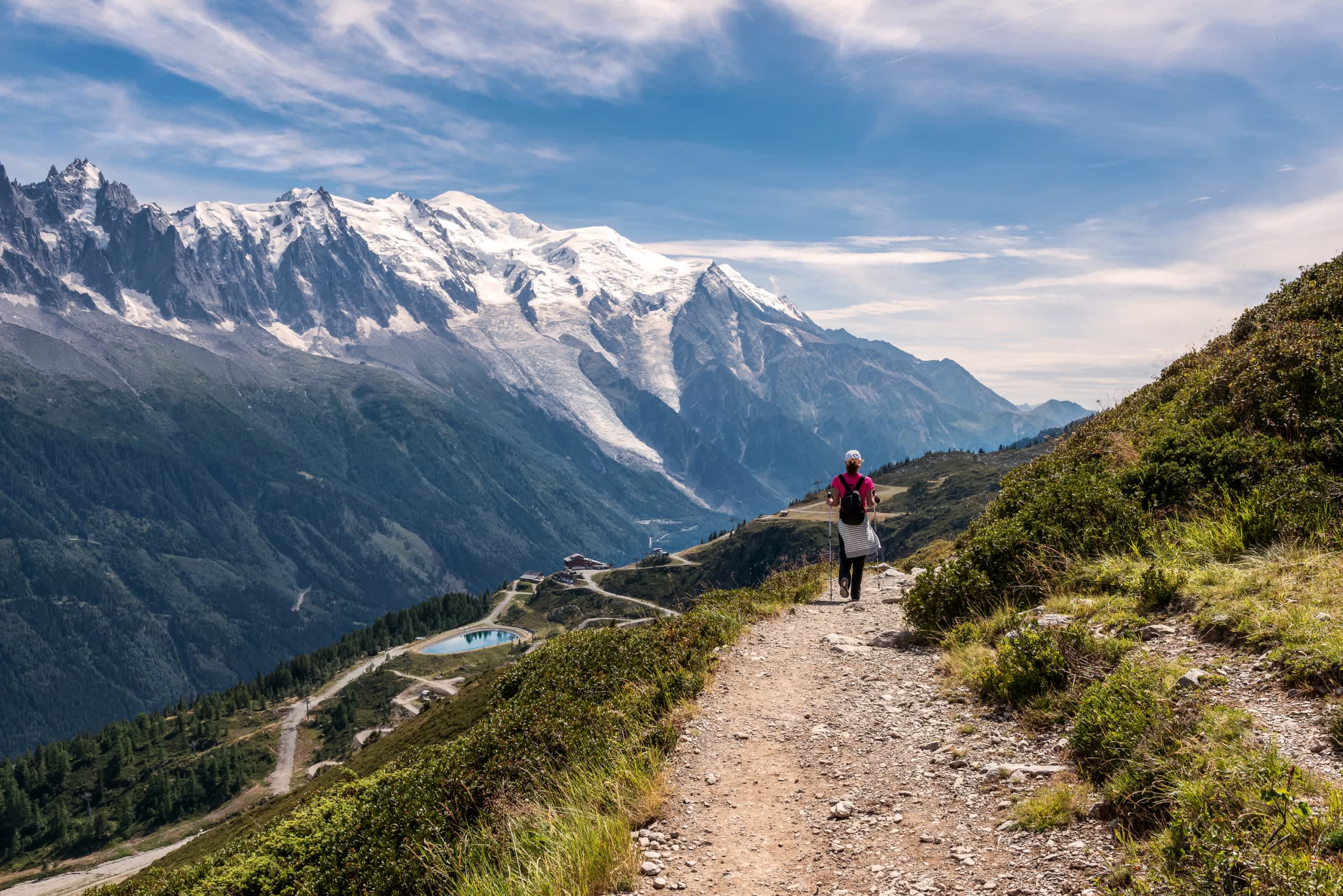 A person hiking on a trail in the foreground, surrounded by majestic snow-capped mountains and a lush, green landscape in the background.