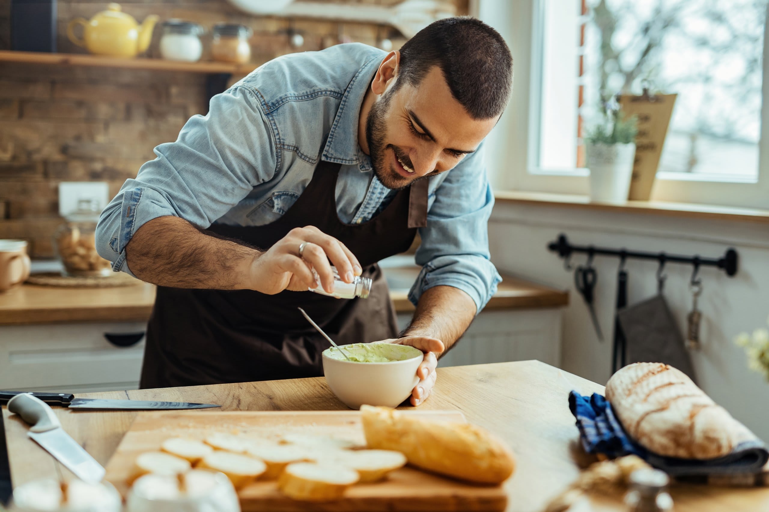 A man in a black apron is carefully preparing food on a wooden surface in a cozy kitchen setting, with various cooking utensils and ingredients visible in the background.