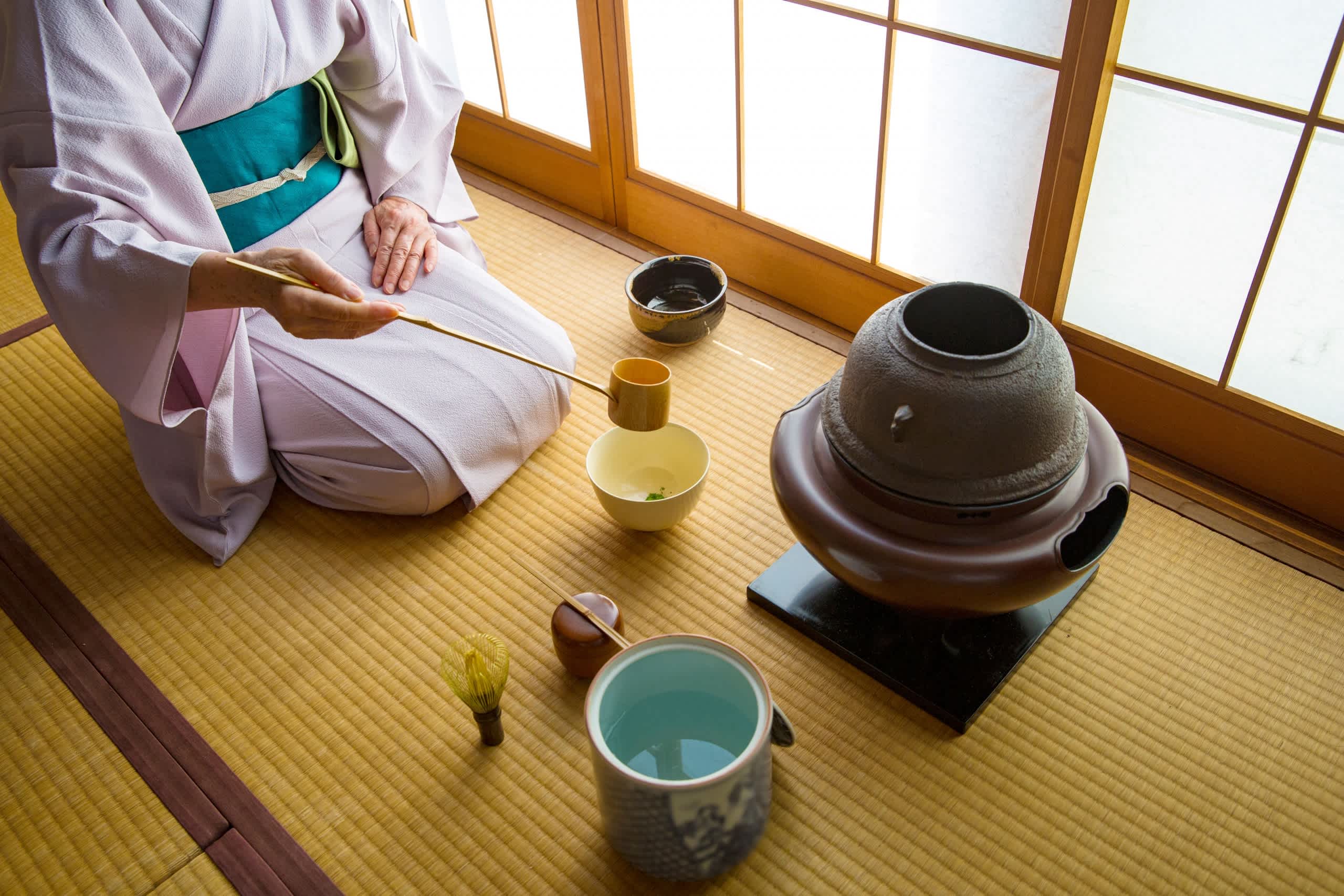 The image depicts a traditional Japanese tea ceremony setting, with a person in a kimono kneeling on a tatami mat floor surrounded by various tea ceremony implements such as ceramic bowls, a tea whisk, and a tea canister.