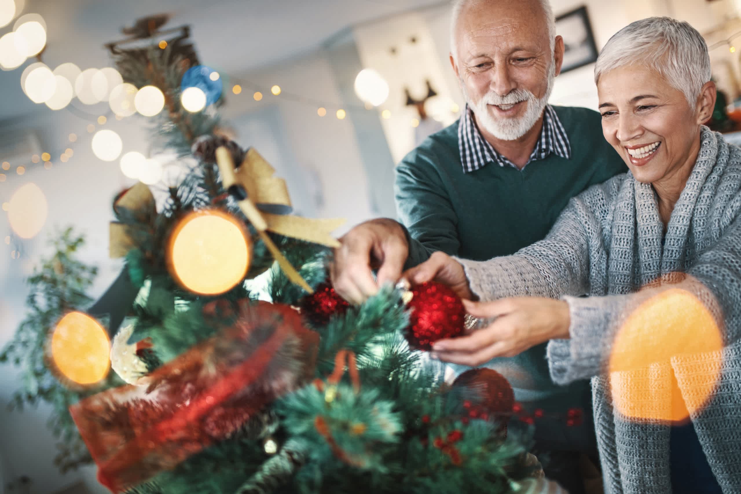 An elderly couple decorating a Christmas tree together, surrounded by festive lights and decorations in the background.