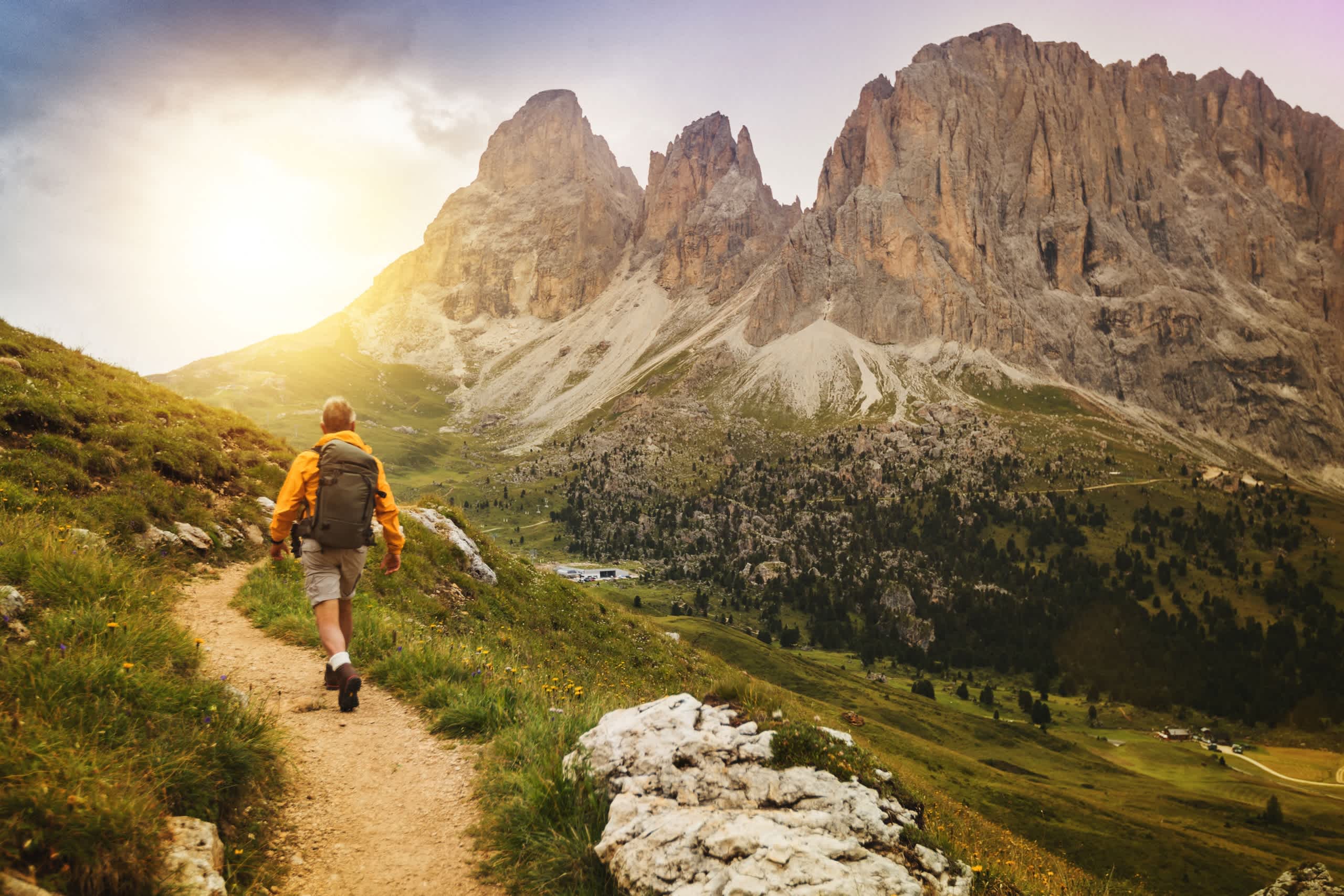 A hiker walks along a trail in a mountainous landscape, with towering, jagged peaks in the background bathed in warm, golden sunlight.