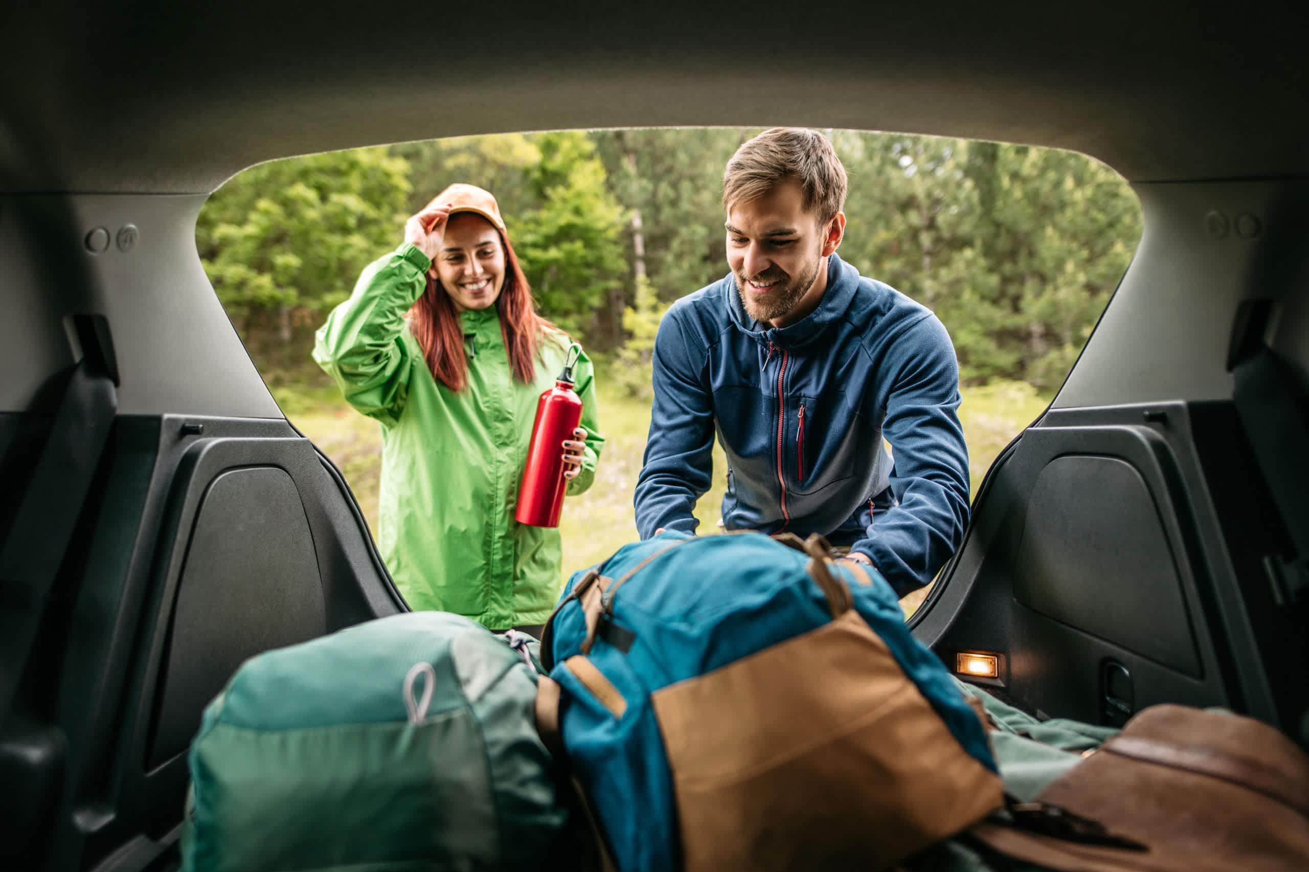 A man and a woman, both wearing outdoor gear, are standing in the back of a vehicle filled with camping equipment and supplies.