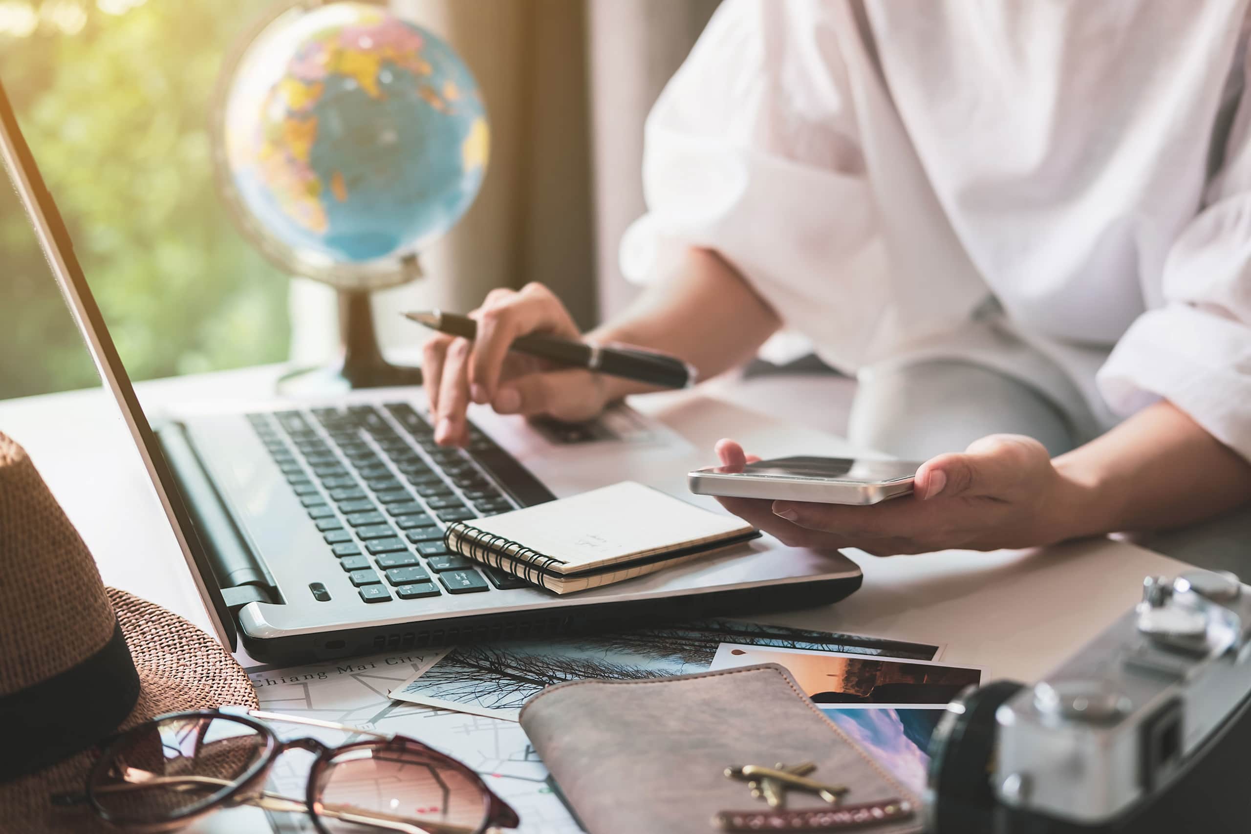 A person's hands are using a laptop and a mobile device on a desk, with a globe and other office supplies visible in the background.
