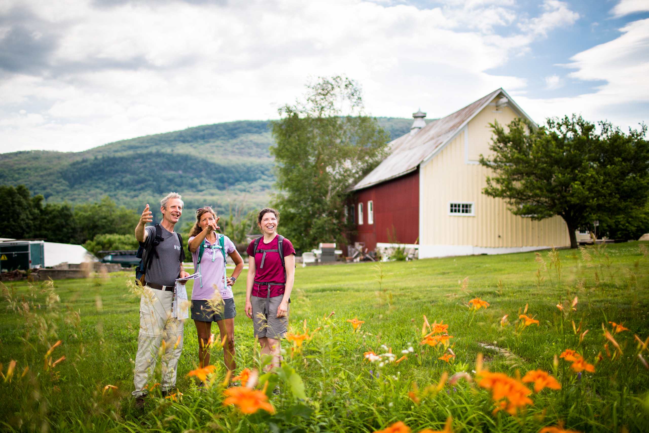 A group of people standing in a grassy field in front of a red barn, with mountains visible in the background.