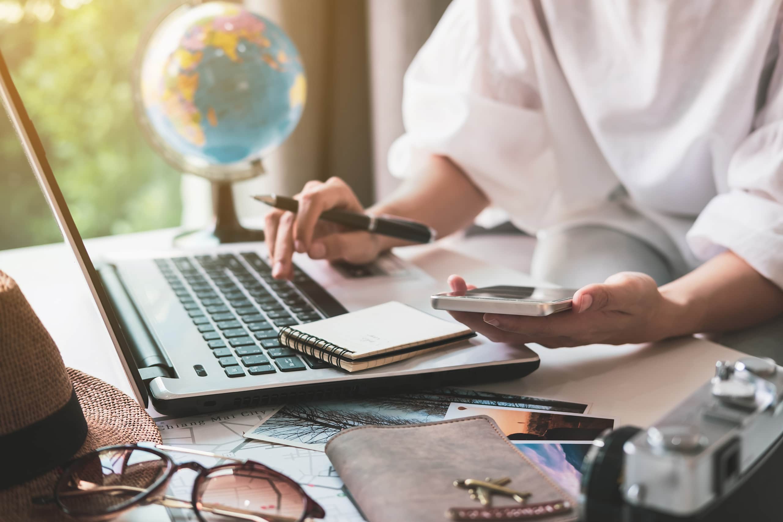 A person's hands are using a laptop and mobile device on a desk, with a globe and other office supplies visible in the background.