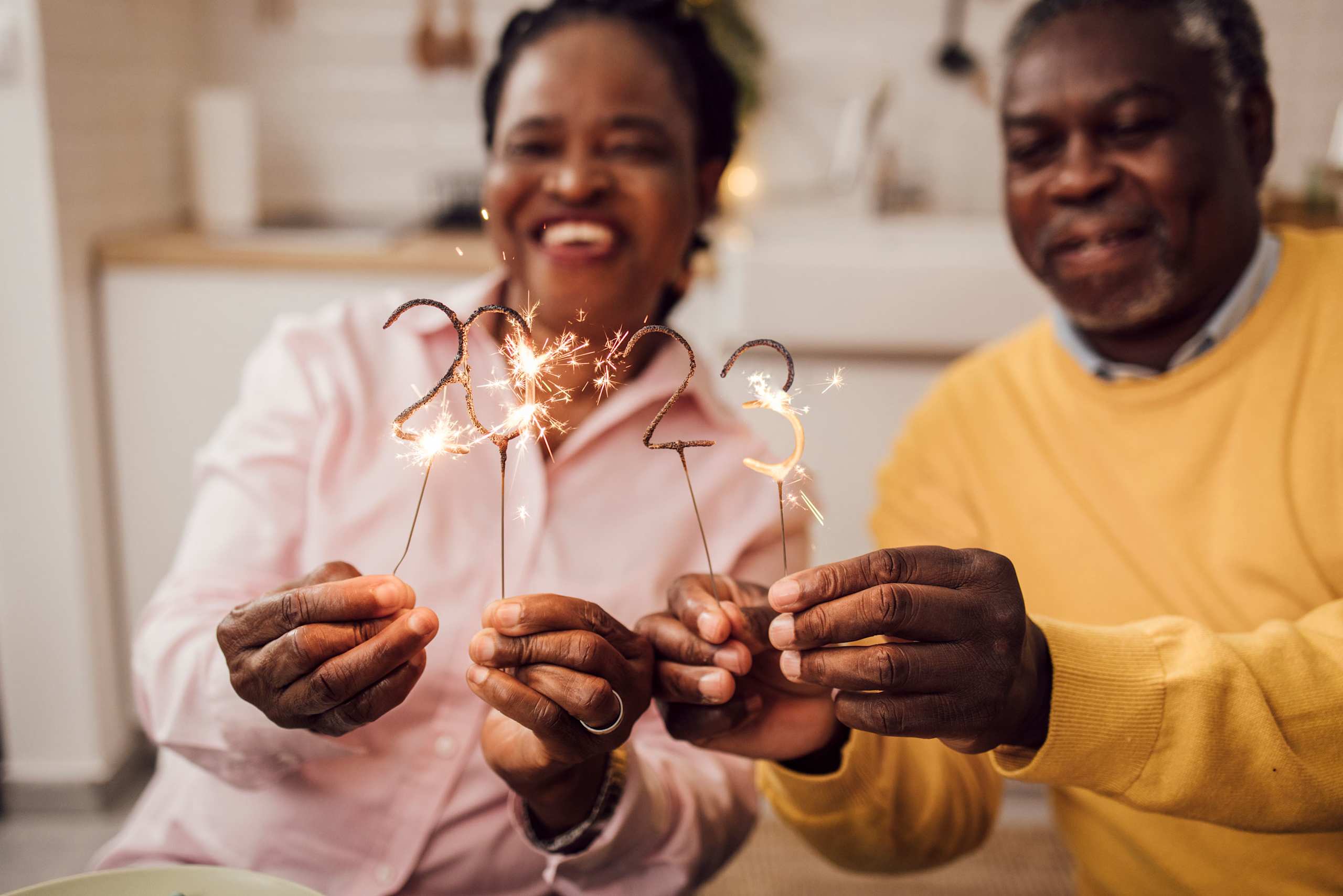 Two elderly individuals, a man and a woman, are holding sparklers and smiling joyfully in what appears to be a cozy indoor setting.