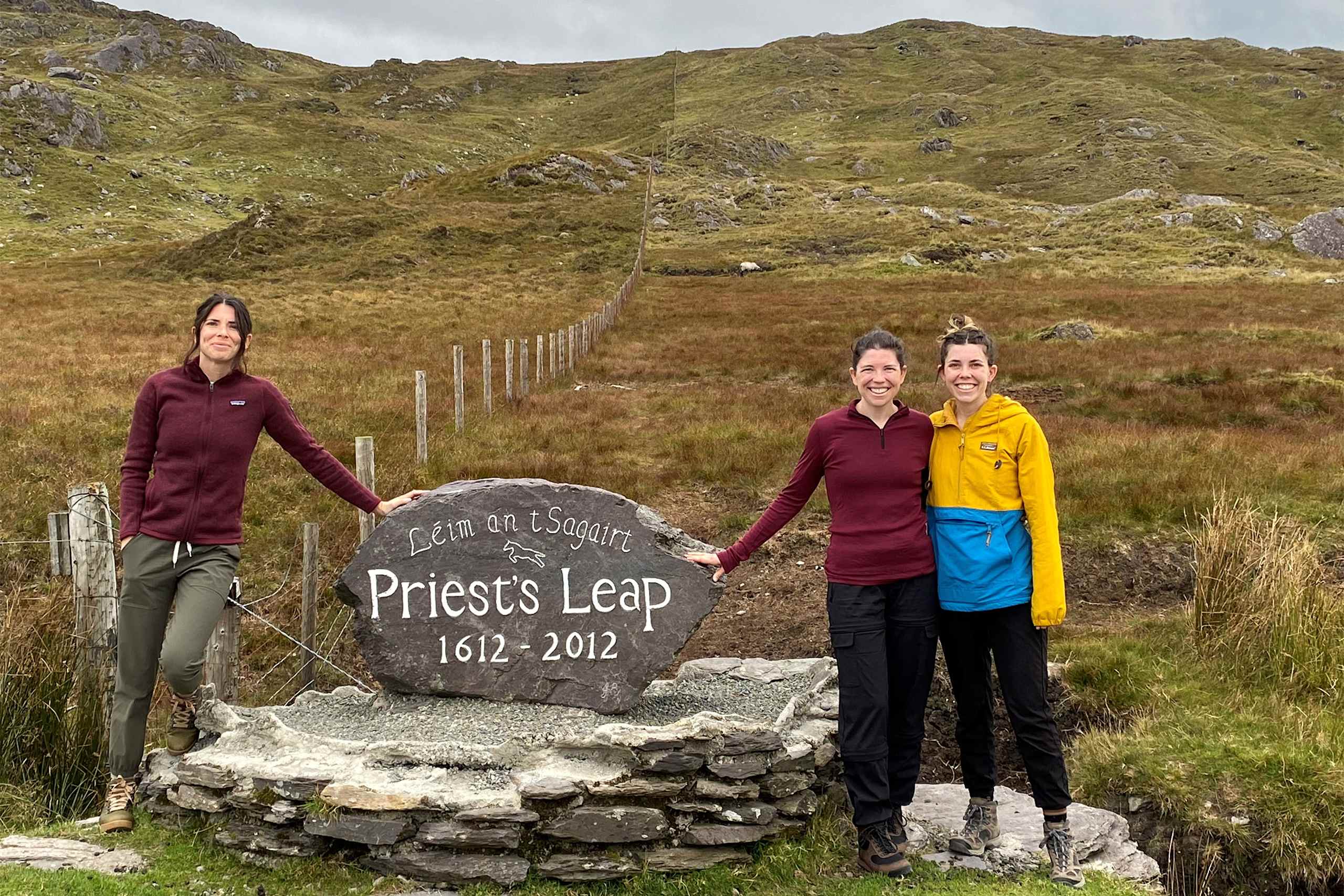 The image shows three women standing in front of a stone marker that commemorates "Priest's Leap" in a rugged, mountainous landscape.