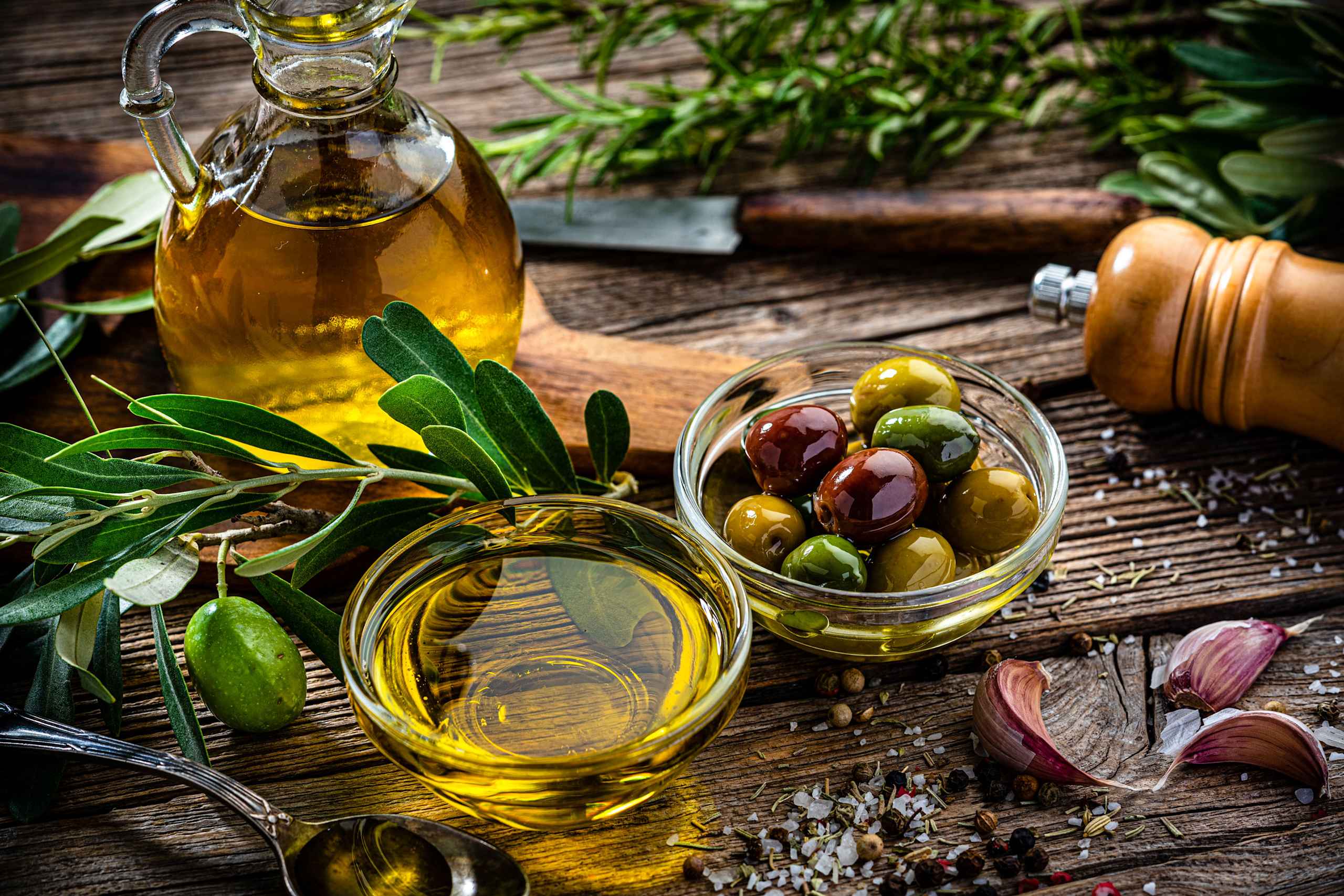 A rustic wooden table with a glass jar filled with green and purple olives, surrounded by bottles of olive oil, fresh herbs, and a mortar and pestle against a backdrop of lush greenery.