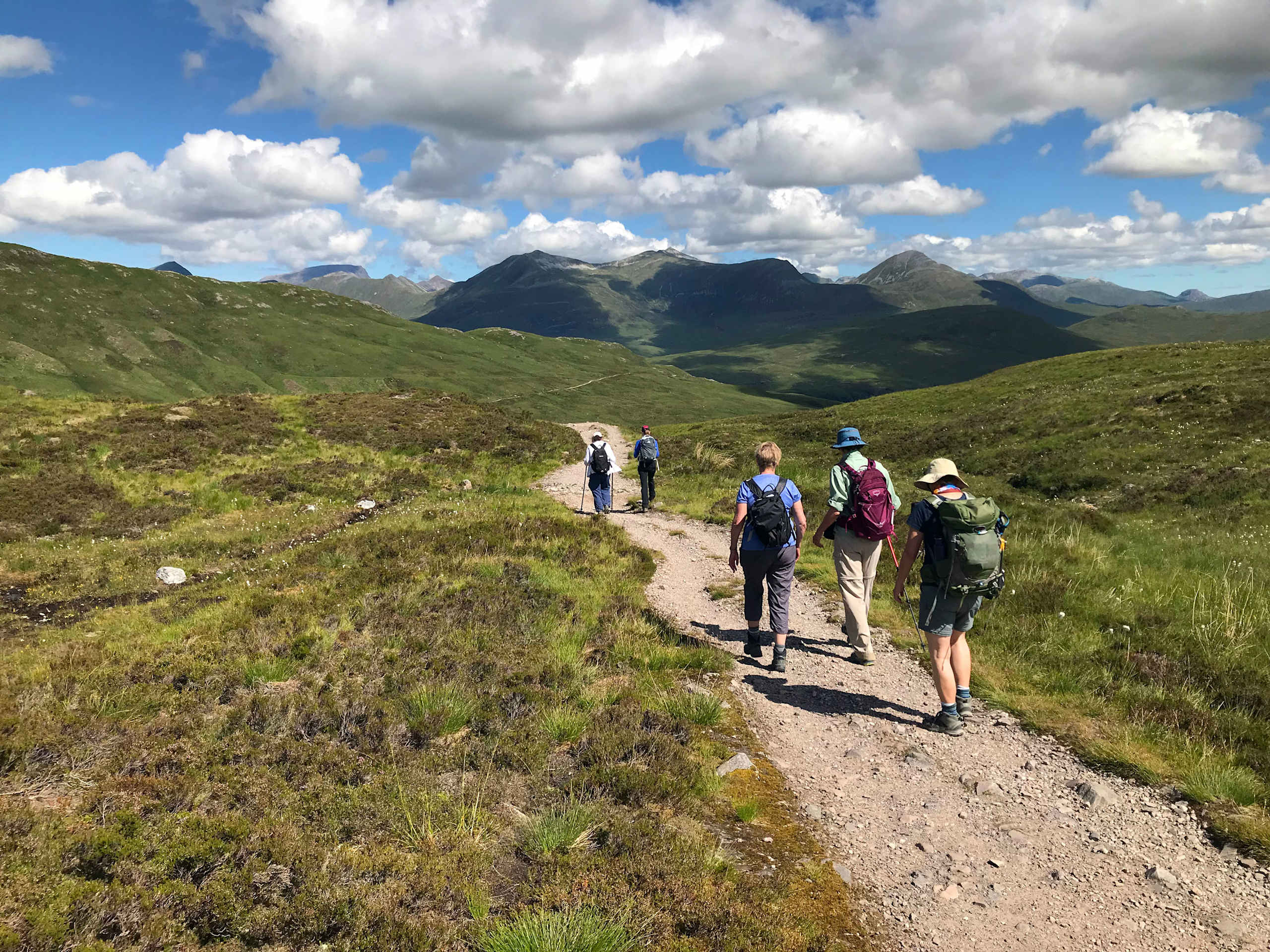 A group of hikers walking along a dirt path through a lush, green landscape with towering mountains in the background under a cloudy blue sky.
