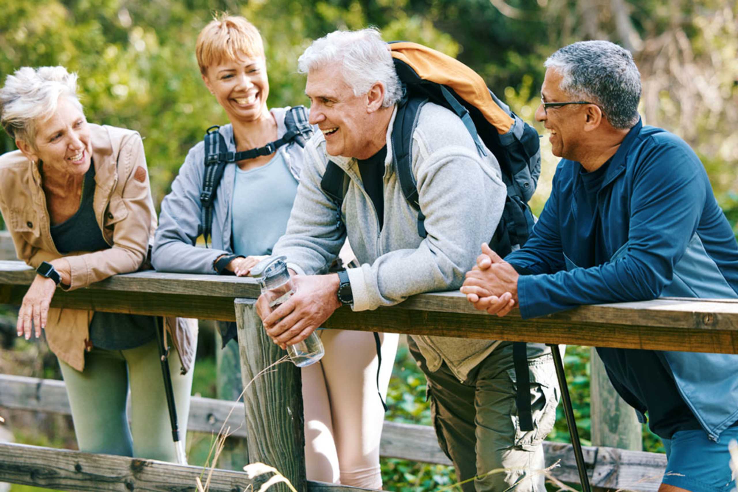 A group of four older adults, two men and two women, are standing on a wooden bridge in a lush, green outdoor setting.