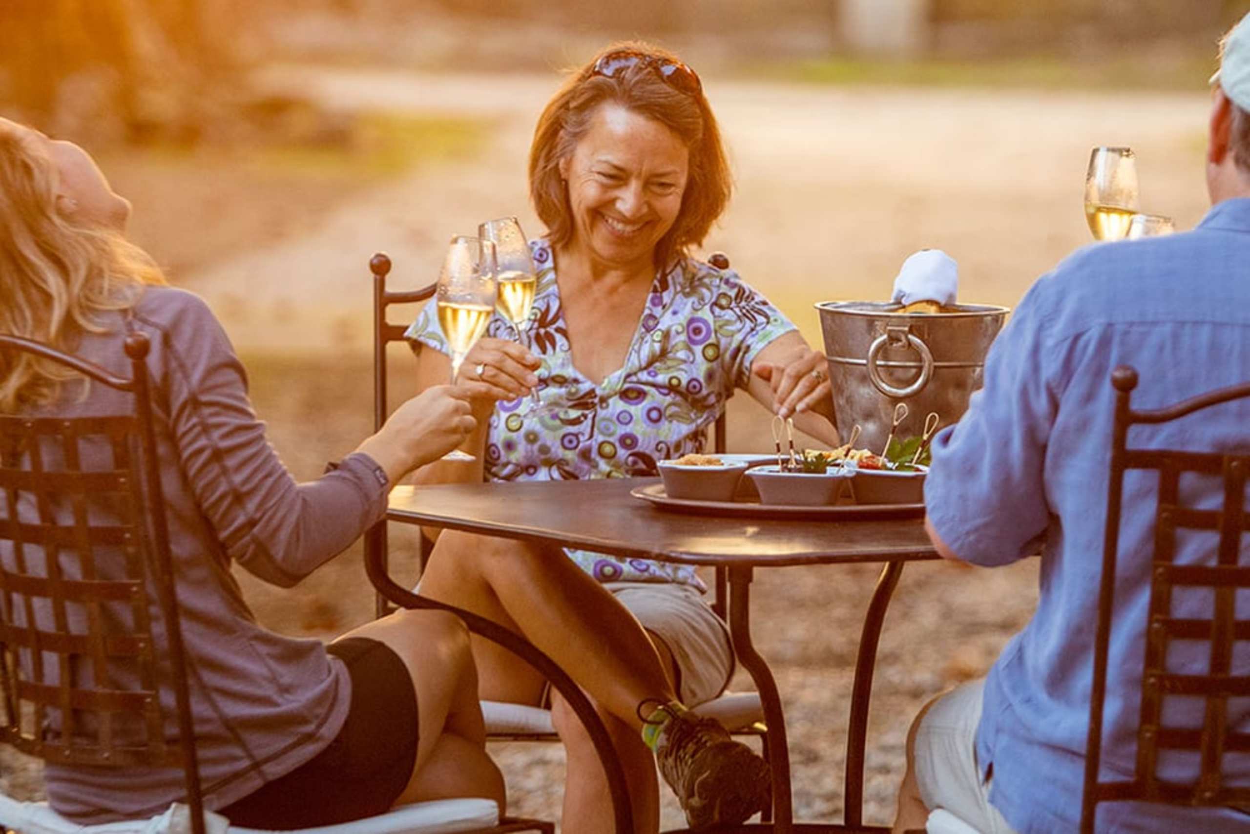 A group of people enjoying a meal outdoors, with a woman in a floral dress sitting at a table and conversing with the others.