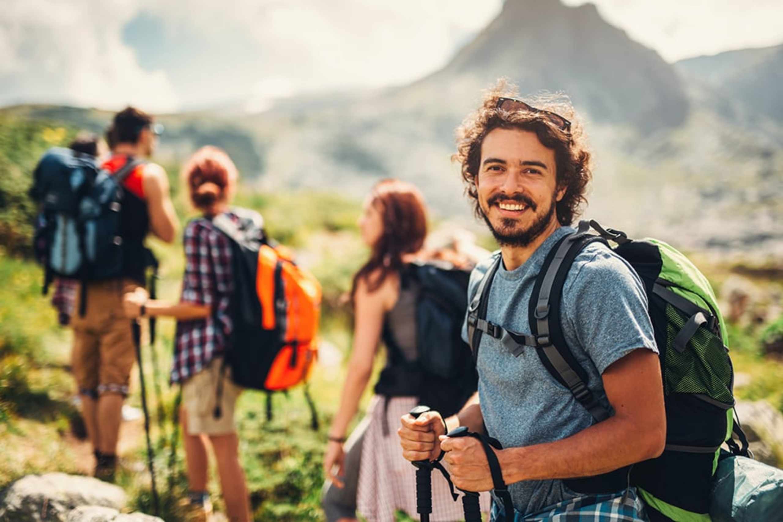 A group of hikers, including a smiling man with a backpack, stand in a scenic mountain landscape with lush vegetation in the background.