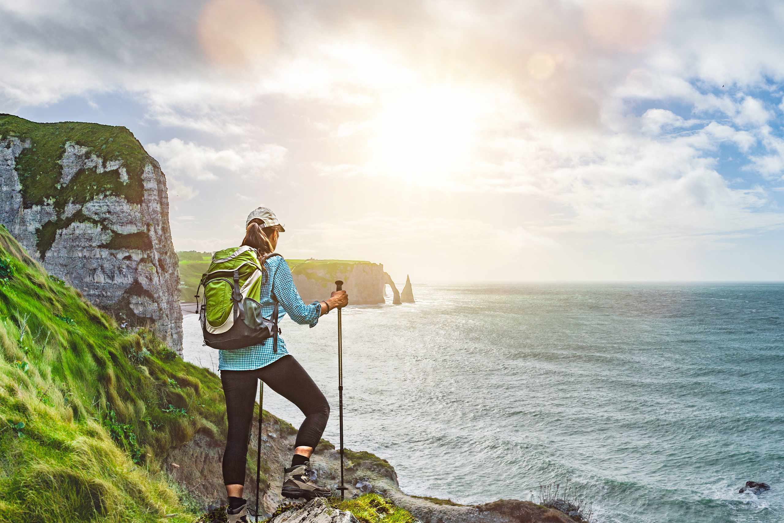 A person with a backpack stands on a rocky cliff overlooking a vast ocean, with dramatic cliffs and a cloudy sky in the background.