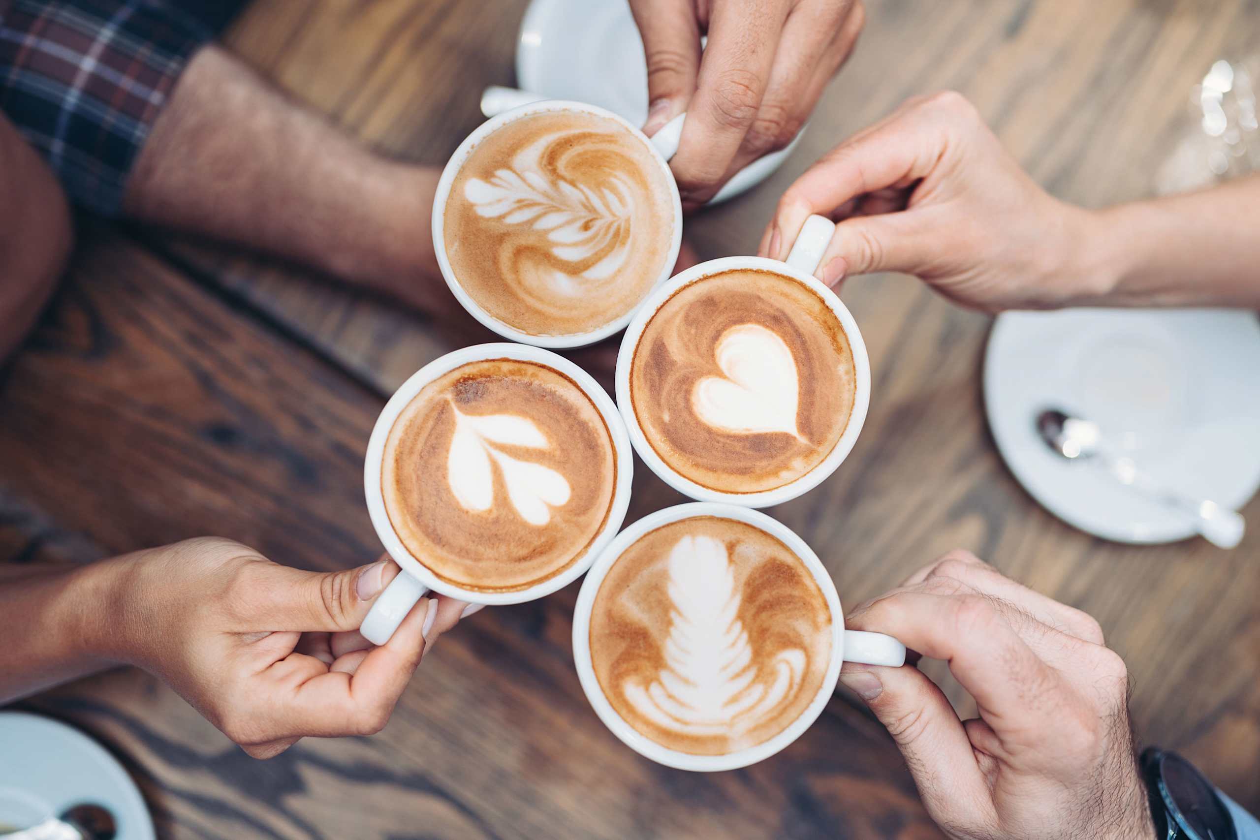Several hands holding cups of coffee with intricate latte art designs, set against a wooden table in a cozy cafe-like setting.