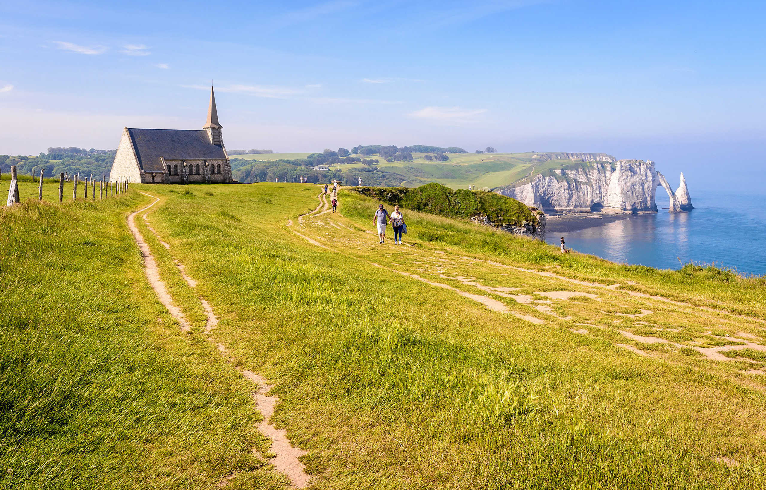 A winding path leads through a grassy field towards a church with a tall spire, with dramatic cliffs and the sea visible in the background.