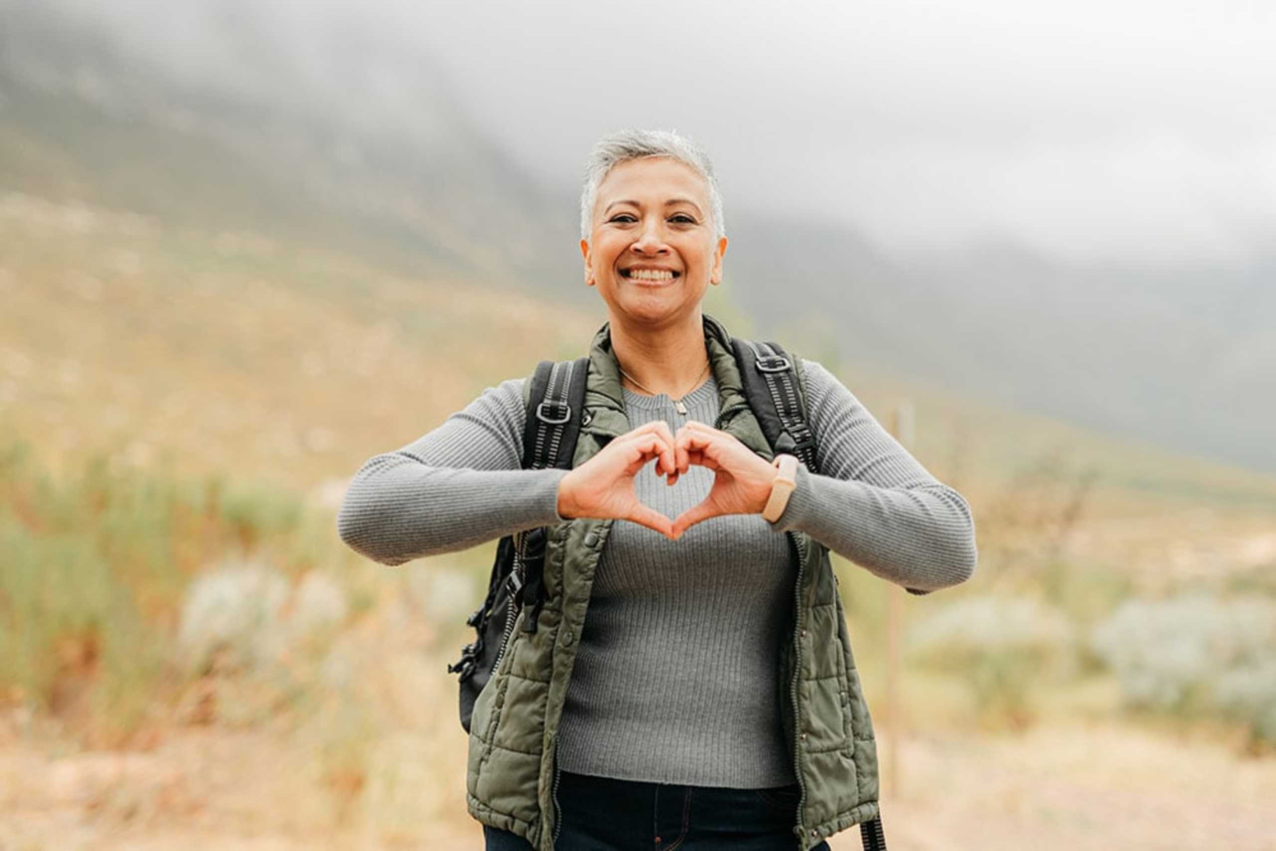 A smiling woman in a gray sweater and vest stands in a grassy field, making a heart shape with her hands against a backdrop of mountains and hazy sky.