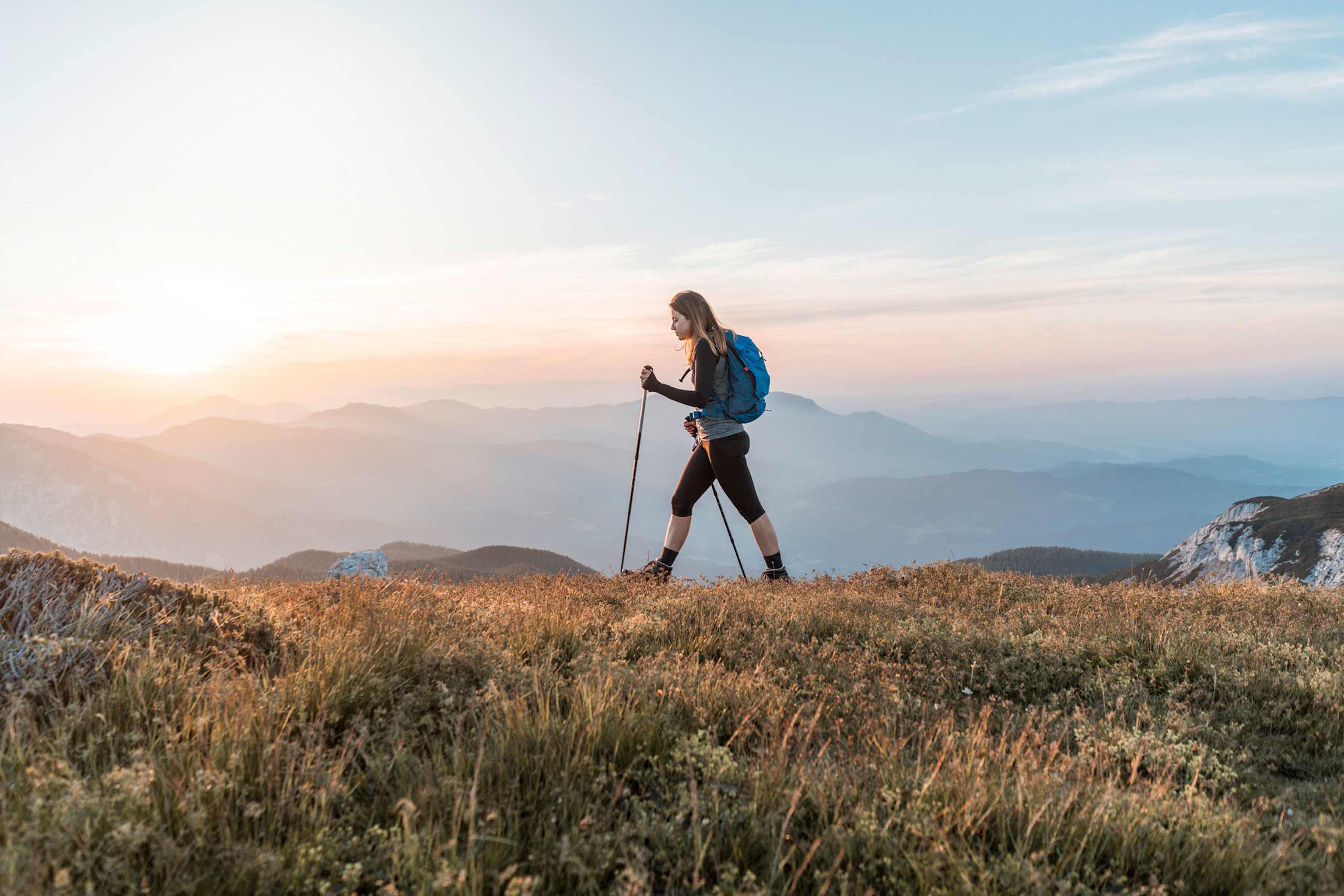 A person with a backpack stands on a grassy hill, surrounded by mountains and a warm, hazy sunset in the background.