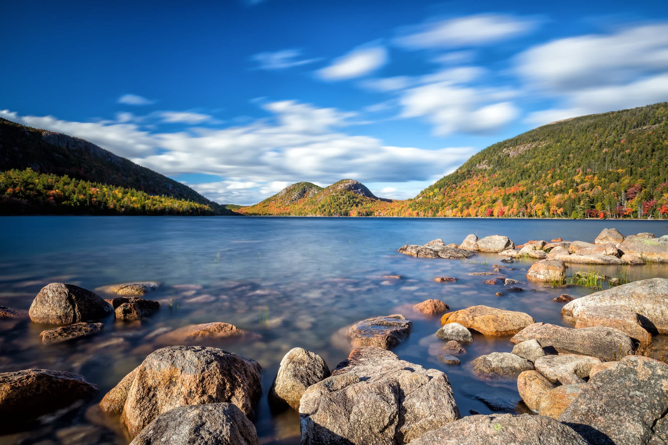 A serene mountain lake surrounded by rocky shores, with lush autumn foliage on the distant hills and a dramatic cloudy sky overhead.