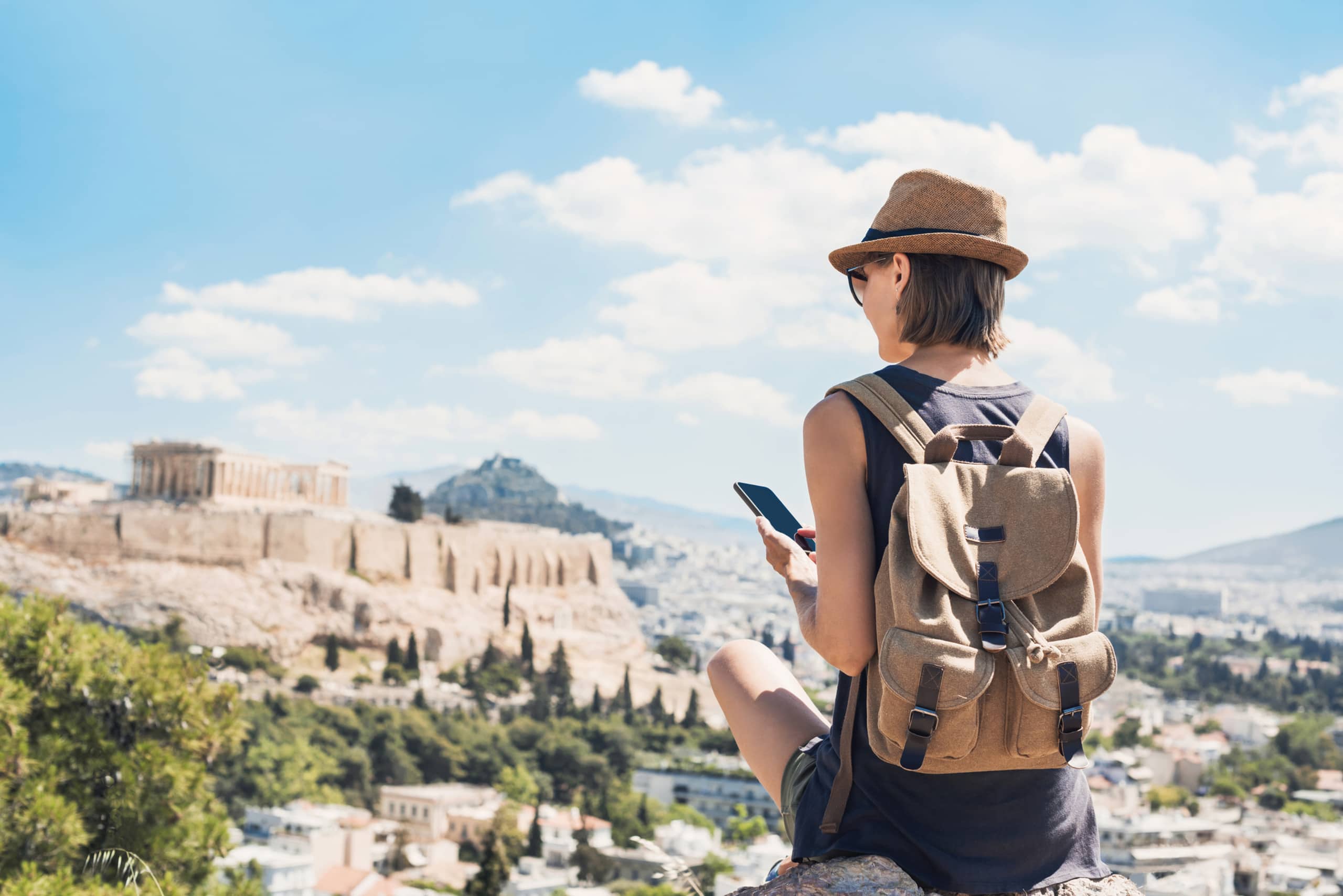 A person wearing a hat and backpack stands facing a scenic view of ancient ruins and buildings against a backdrop of cloudy blue sky.