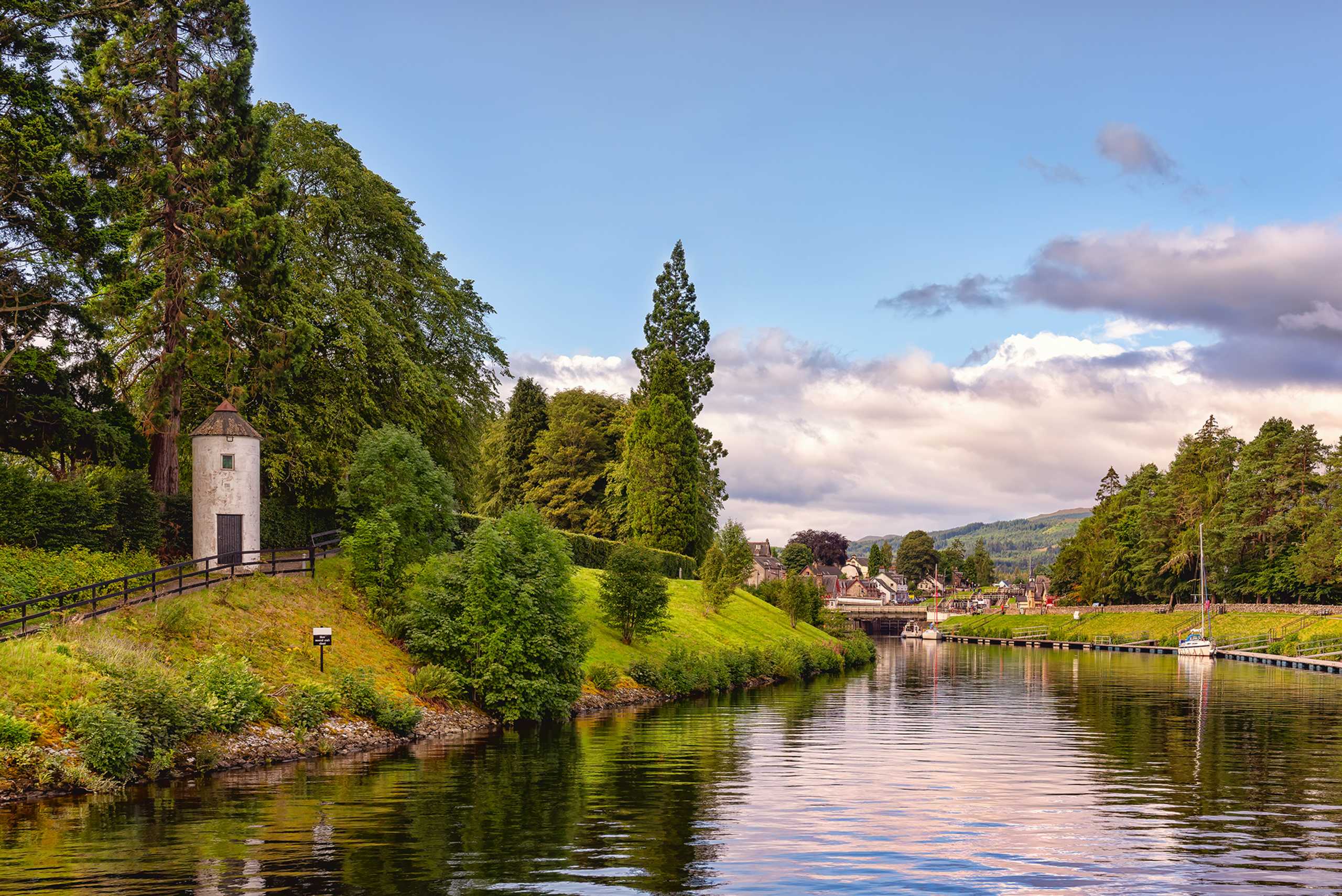 A serene canal surrounded by lush greenery, with a small tower structure visible in the distance, reflecting in the calm waters.