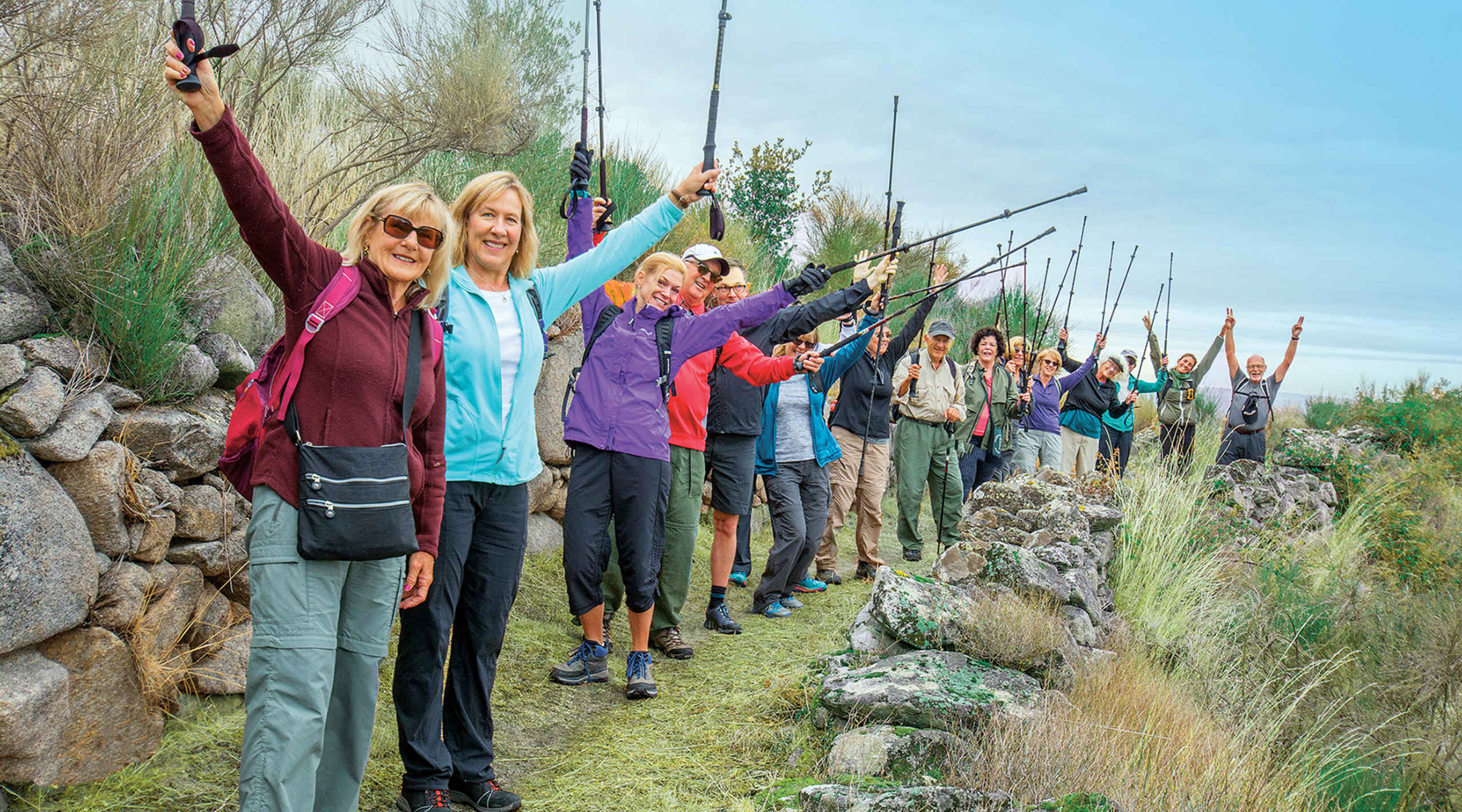 A group of people, dressed in colorful outdoor attire, are standing together on a rocky trail surrounded by lush vegetation and a clear blue sky.