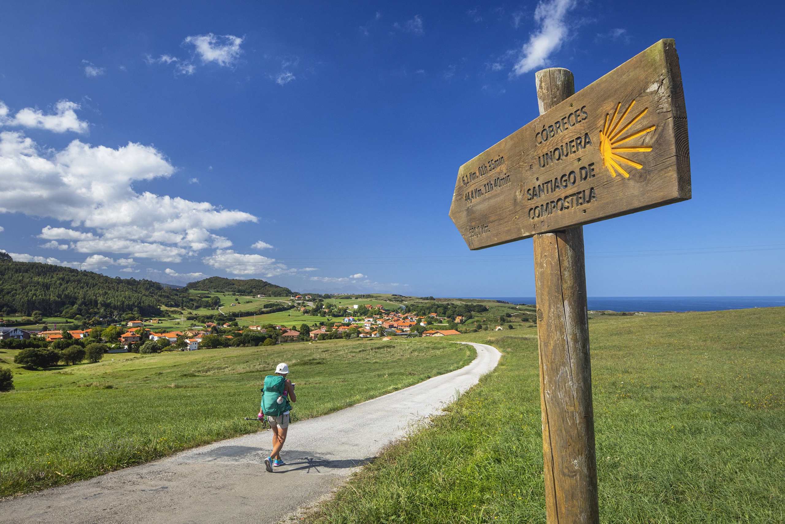 A wooden signpost stands in a grassy field, pointing the way along a winding path that leads to a picturesque village nestled among rolling hills and a distant coastline under a bright, cloudy sky.