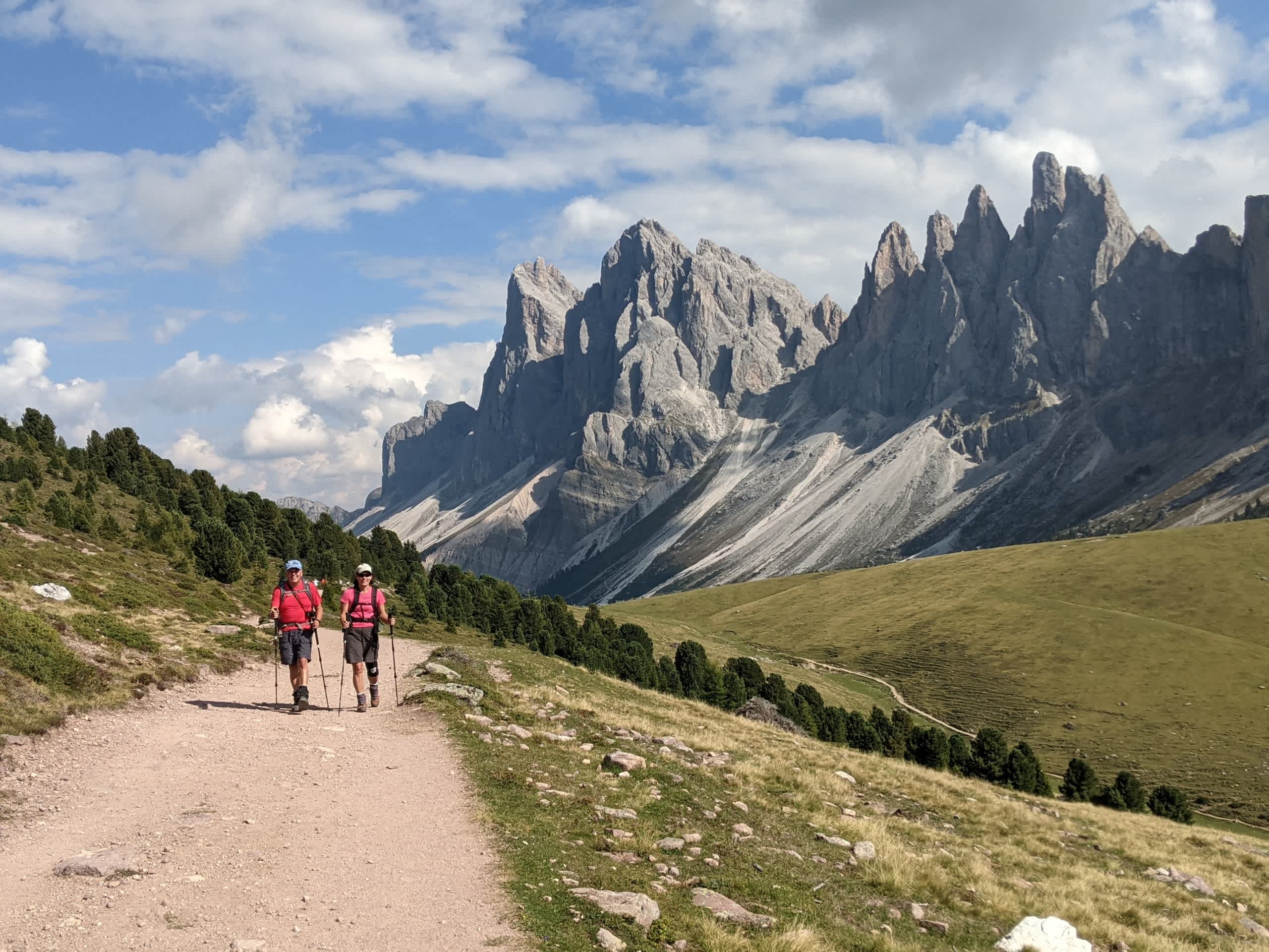 The image depicts a scenic mountain landscape with rugged, snow-capped peaks in the background, and two hikers walking along a dirt path in the foreground.