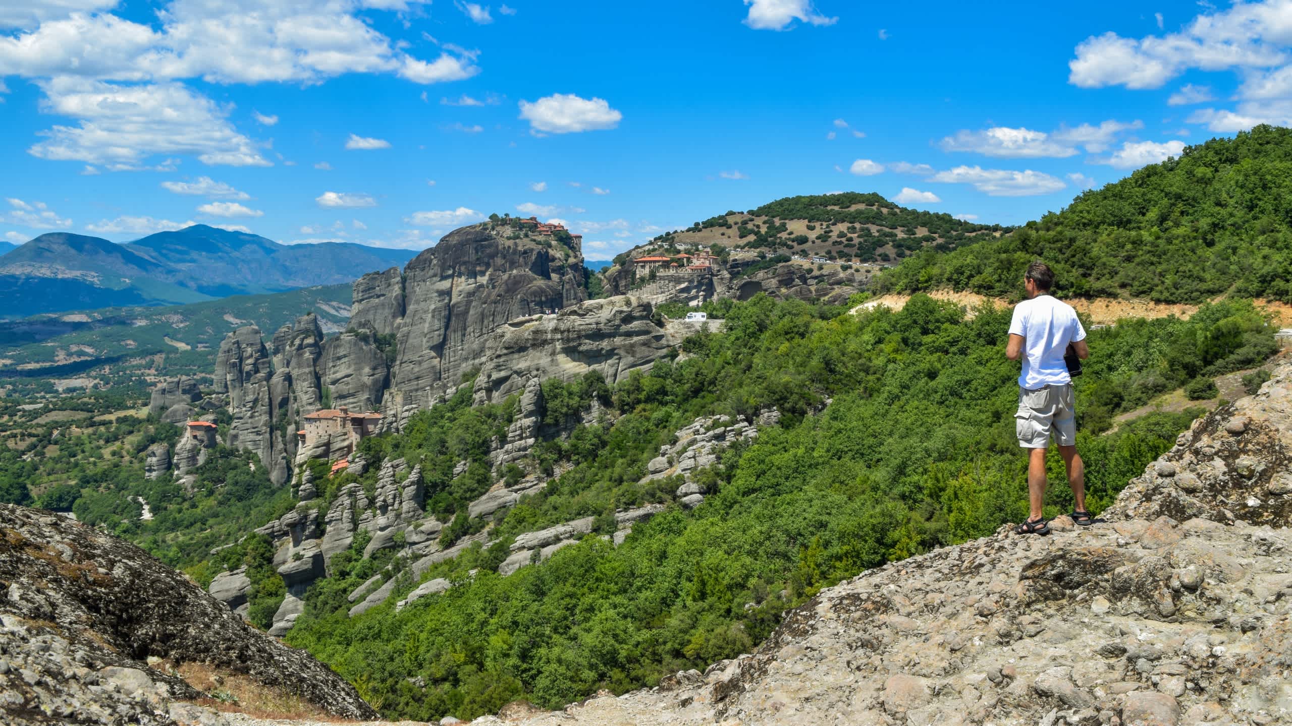 A person stands on a rocky cliff overlooking a lush, forested landscape with towering mountains in the distance under a bright, cloudy sky.