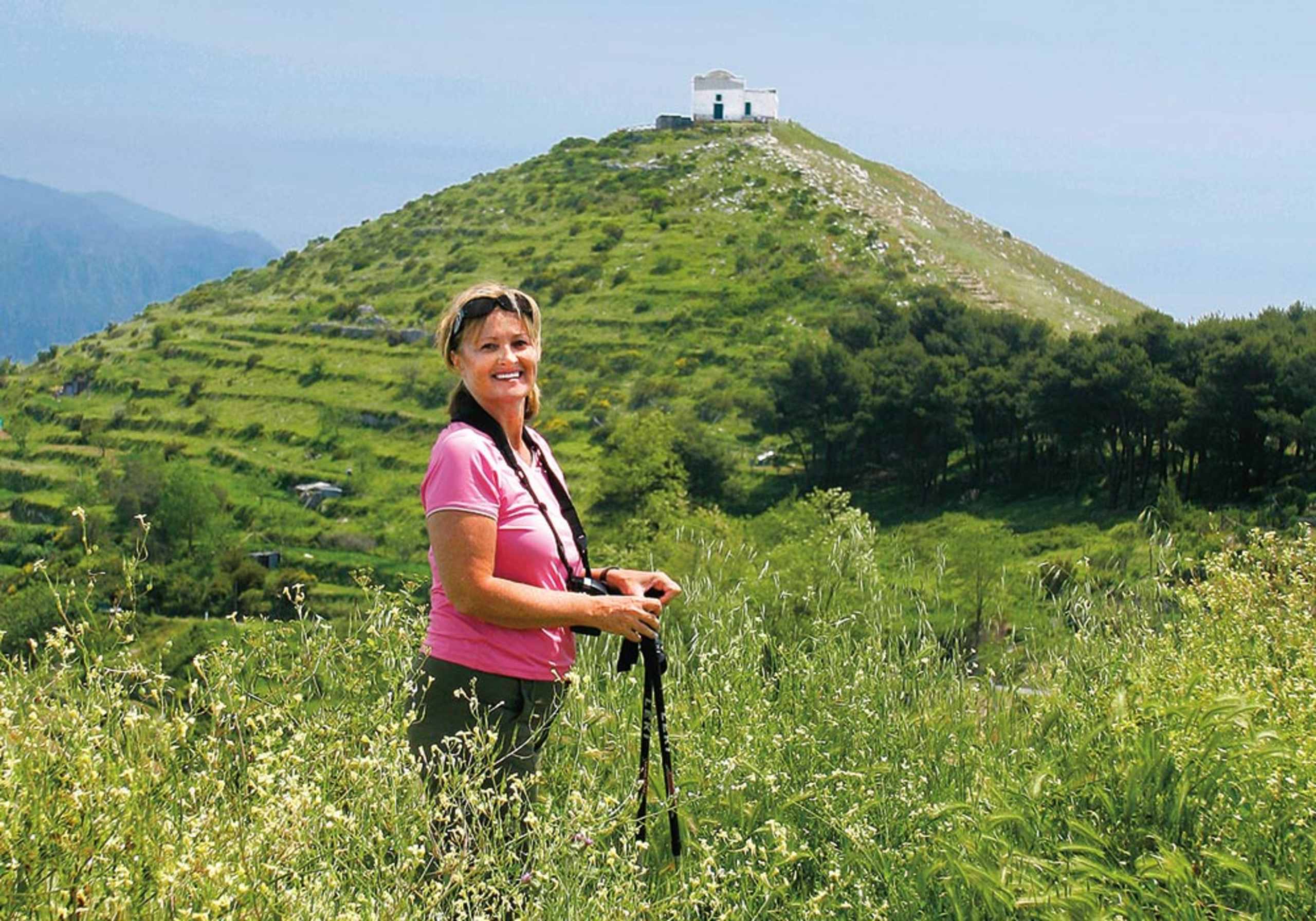 A woman in a pink shirt stands in a grassy field, with a hill topped by a small building visible in the background.