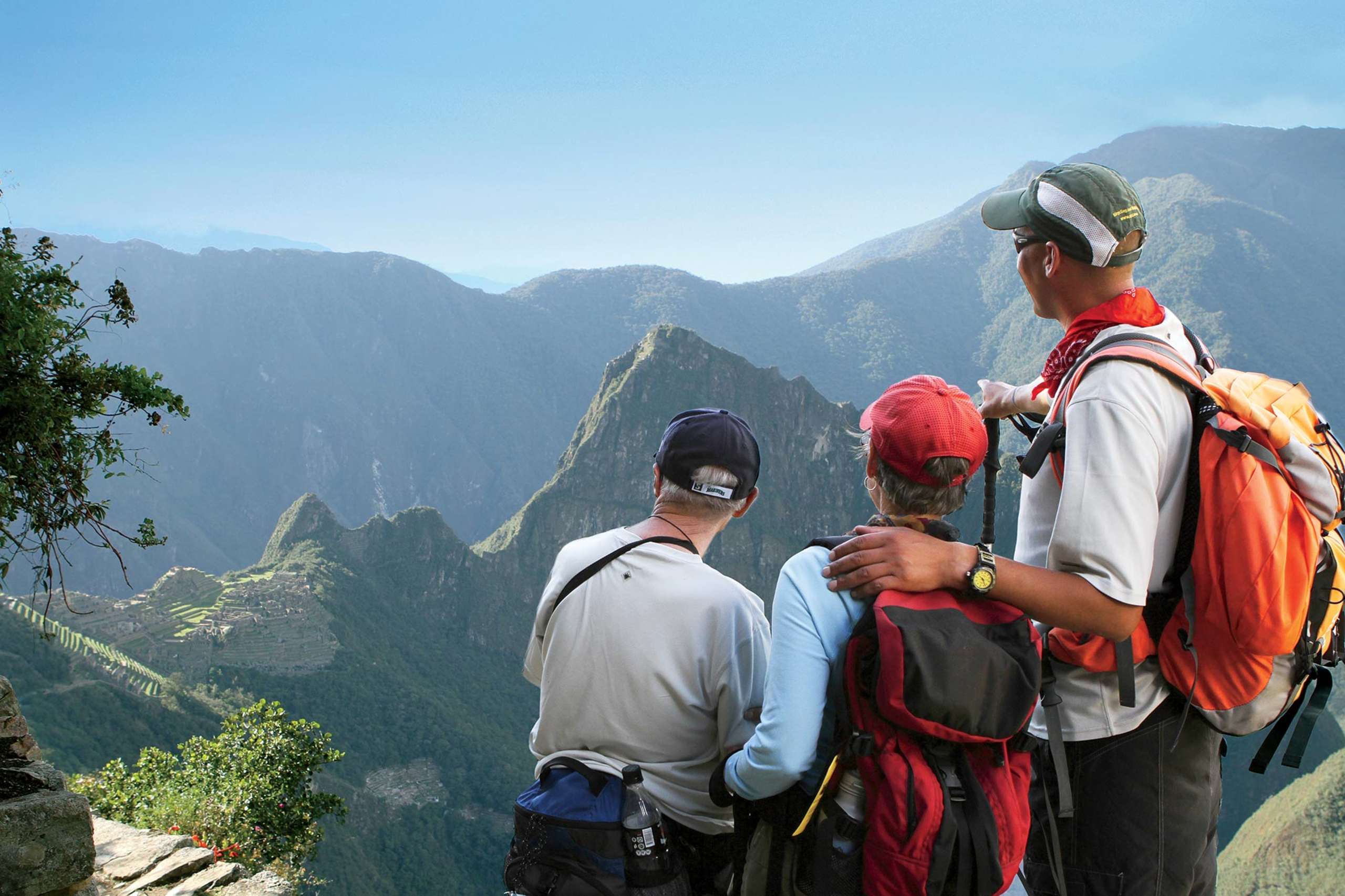 Two hikers stand on a mountain trail, gazing out at the stunning landscape of rugged peaks and lush valleys in the distance.