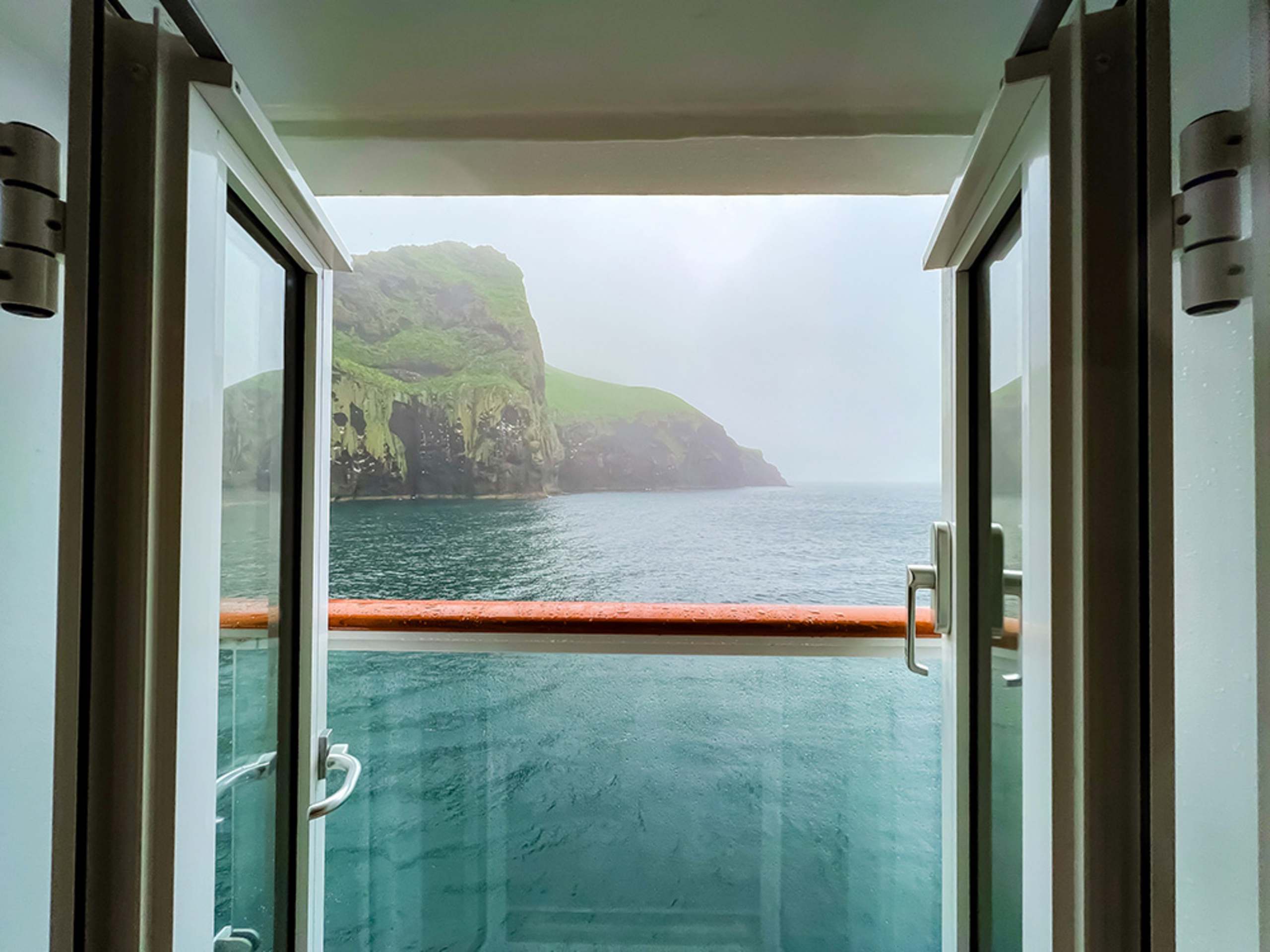 A glass-enclosed balcony overlooking a serene, misty lake surrounded by towering cliffs in the background.