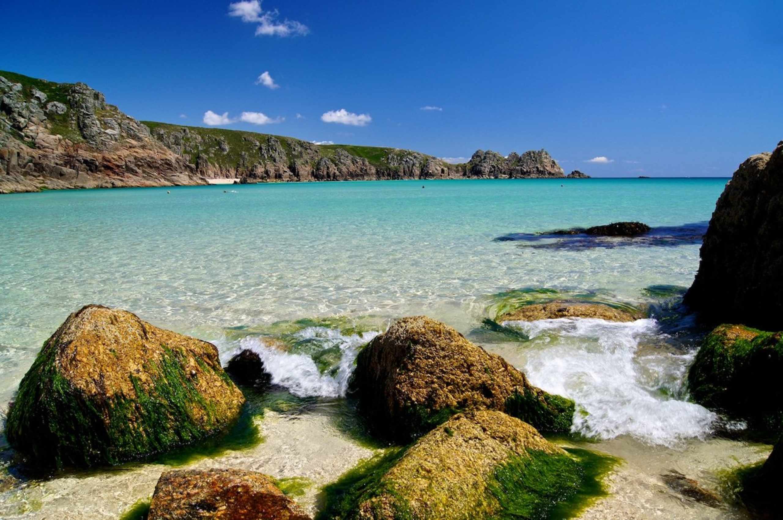 A stunning turquoise bay surrounded by rugged cliffs, with waves crashing against the mossy rocks in the foreground.