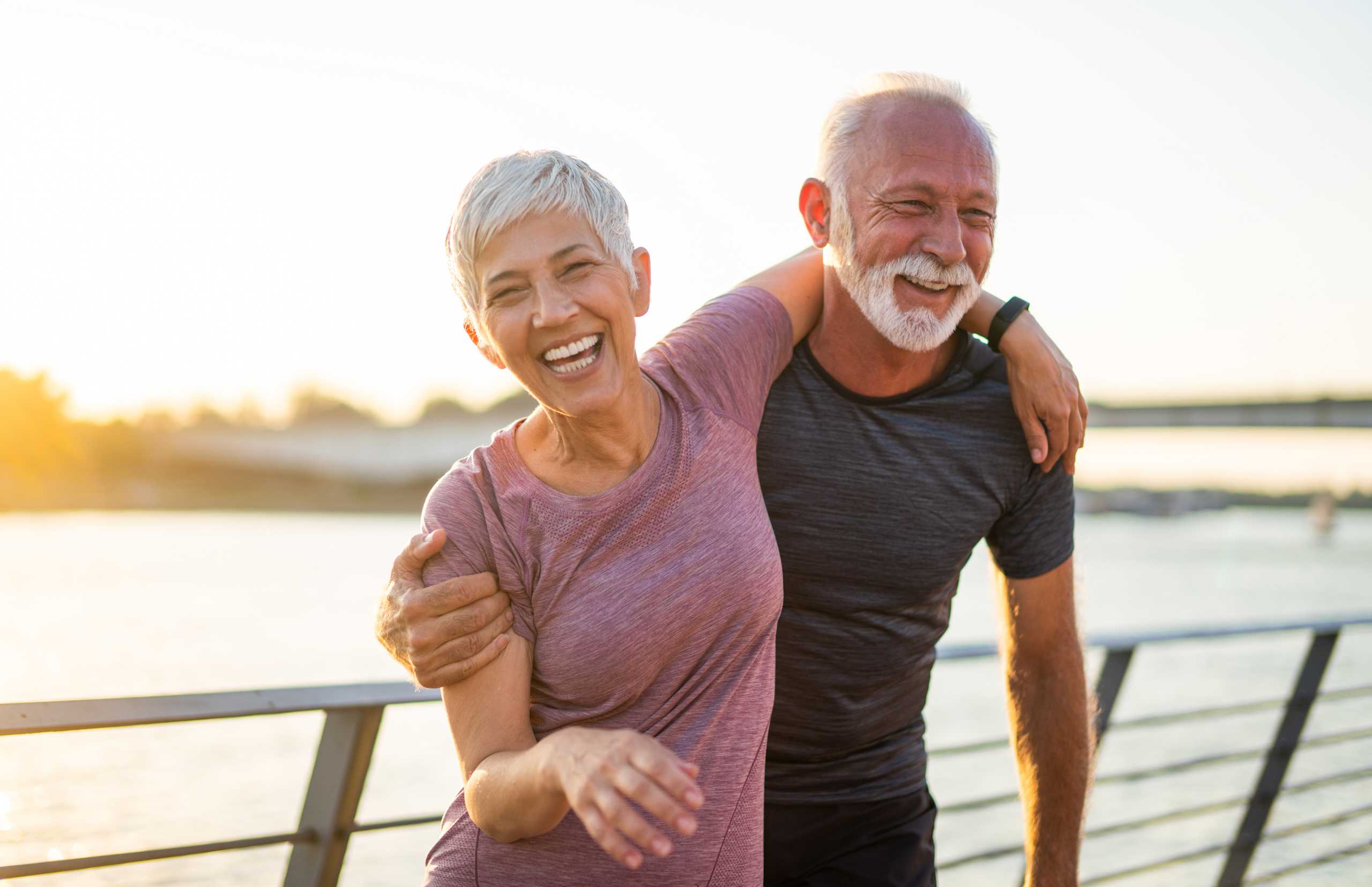 A joyful couple laughs together by the water at sunset, walking arm-in-arm along a railing with warm golden light behind them.