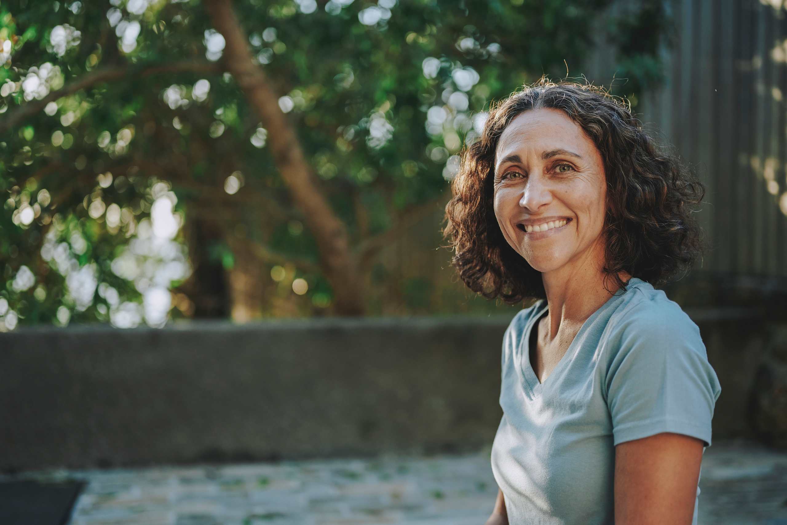 A woman smiles warmly outdoors, sunlight filtering through leafy trees behind her on a quiet stone path.