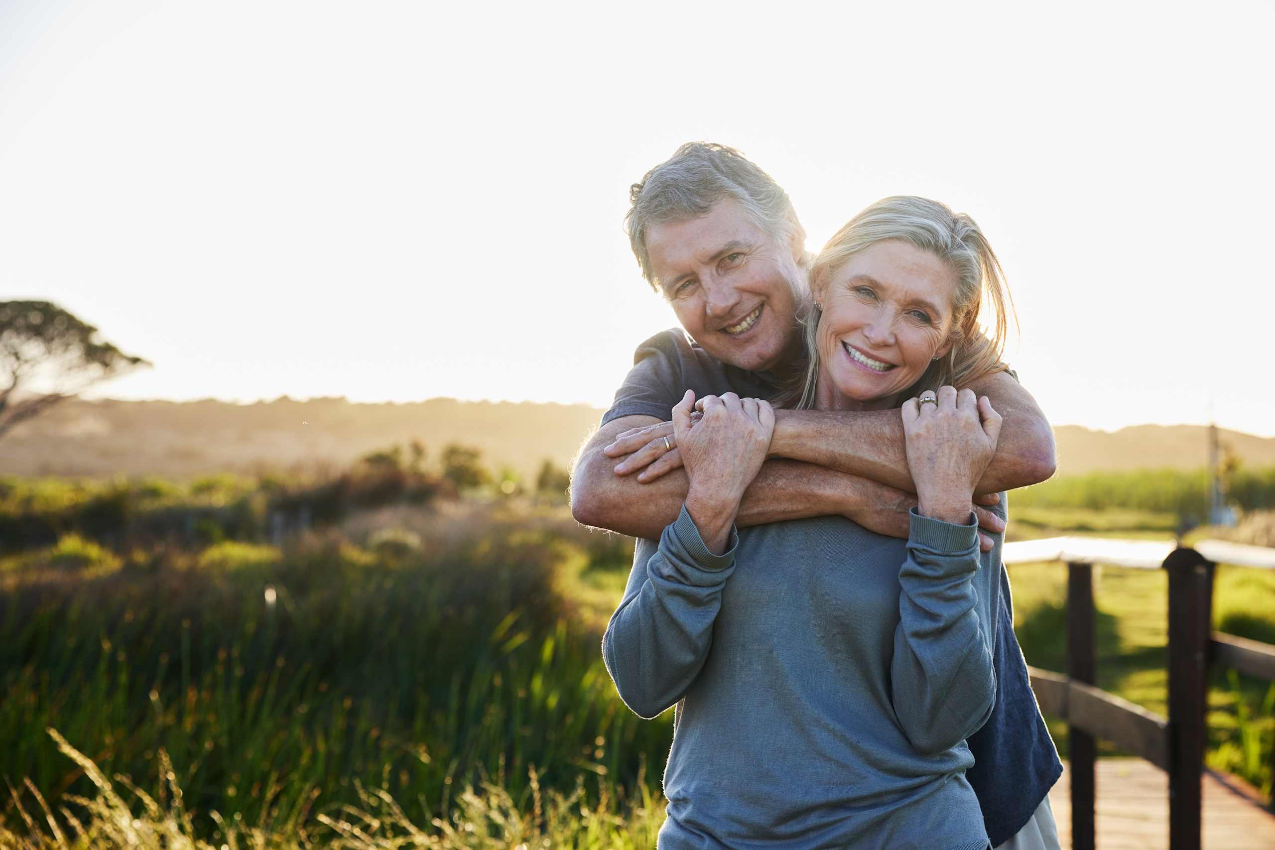 A couple shares a warm embrace at sunset on a quiet boardwalk, smiling as golden light falls over the surrounding wetlands.