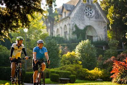 Two cyclists riding their bicycles on a path surrounded by lush greenery and a picturesque church in the background.