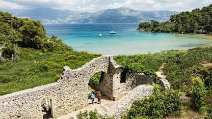 Two travelers explore ancient stone ruins overlooking a turquoise bay in Croatia, with forested hills and sailboats in the distance.