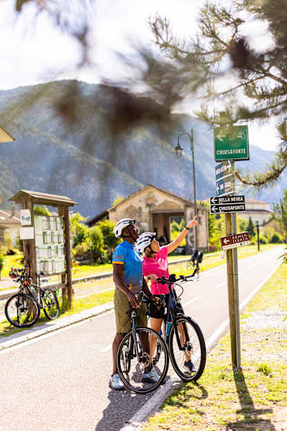 The image shows a couple riding bicycles on a path surrounded by trees and buildings, with a sign indicating a nearby community center.