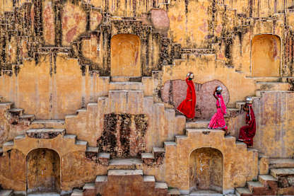 The image depicts a grand, ornate architectural structure with intricate details and patterns, set against a backdrop of vibrant colors and textures. In the foreground, two figures in colorful, flowing robes stand amidst the elaborate surroundings, adding a sense of scale and human presence to the scene.