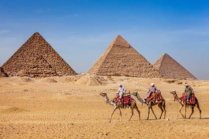 The image depicts three people riding camels in the foreground, with the iconic pyramids of Giza visible in the background against a clear blue sky.